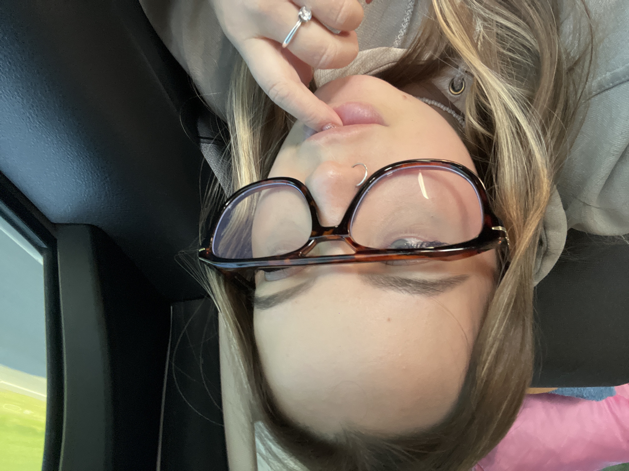 An upside-down close-up photo shows a person, likely female, with light brown or blonde hair wearing glasses with tortoise-shell frames. A hand, with a silver ring featuring a clear stone on one finger, is gently touching the person's lips. A small hoop piercing is visible on the nose. The person is wearing a light-colored, possibly grey or light green, hooded top.
The setting appears to be inside a car, indicated by dark interior elements like the headliner and window frame visible around the person. Through the car window on the left, a bright green field and a light blue sky are visible, suggesting it is daytime with clear weather. A pink object, possibly a pillow or blanket, is partially visible in the bottom right corner of the frame. The overall image captures a relaxed moment inside a vehicle, possibly while traveling. The location context is Ternovskaya, Russia. No visible text is present.