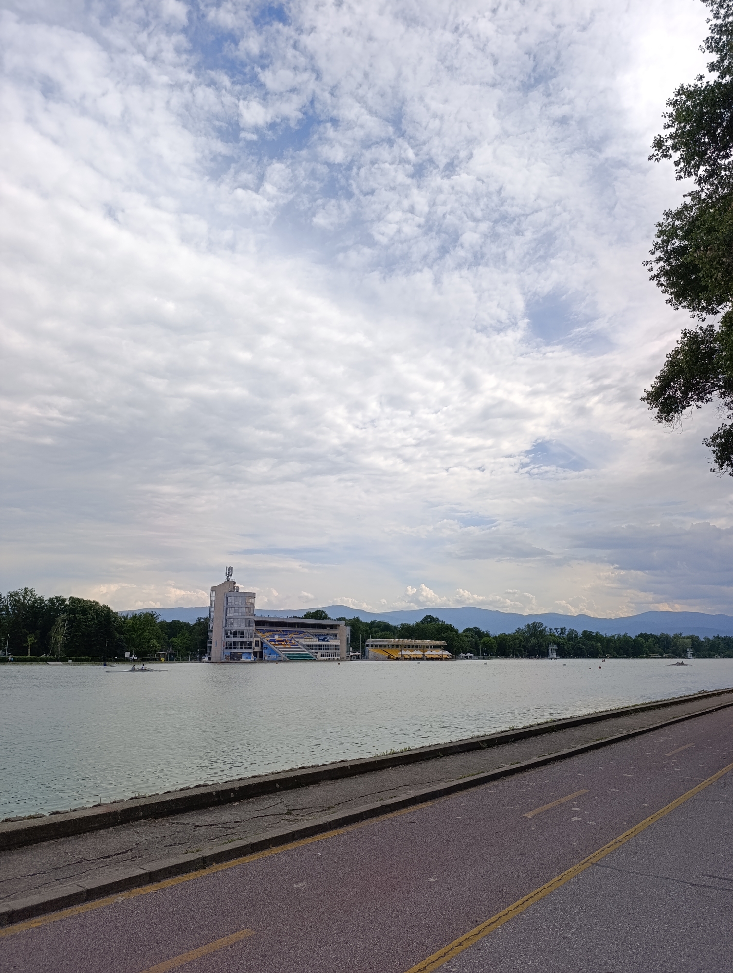 The image presents a wide view of a rowing canal in Plovdiv, Bulgaria, under a cloudy sky. In the foreground, a dark gray paved road with double yellow lines runs alongside the water, separated by a concrete edge. The canal water is calm with a light green or murky appearance.
Across the canal, a prominent multi-story building with a tower-like structure and spectator seating is visible, characteristic of a rowing center or grandstand. An antenna is mounted on its roof. To its left, a longer, low-rise building with a yellow roof can be seen further down the bank, possibly a boathouse or recreational facility.
Two individuals are actively rowing a white shell boat on the canal in front of the main building, indicating ongoing training or competition. The banks of the canal are lined with dense green trees, and in the distance, hazy mountains complete the backdrop. The sky is mostly covered by white and gray clouds, with occasional glimpses of blue sky. A large tree with dark foliage partially frames the scene from the right side. The overall atmosphere suggests a daytime scene, possibly late morning or afternoon, with functional sporting facilities set within a natural landscape. No legible text is visible in the image.