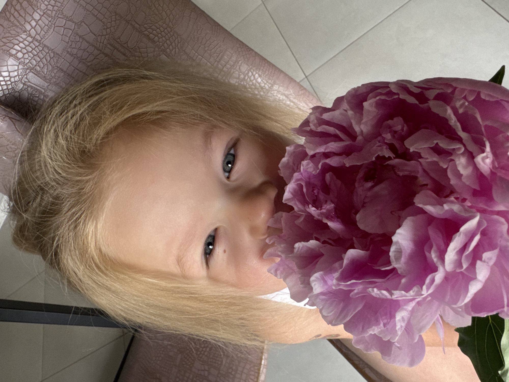 A young child with blonde hair and blue eyes is depicted in a close-up photograph, holding a large, vibrant pink peony flower. The flower is positioned to cover the child's nose and mouth area. The child's blue eyes are directed towards the camera, showing a slight crinkle at the corners. A small mole is visible below the child's left eye. The background on the upper right features a surface with a raised, reptilian-skin-like texture in a brownish-pink hue, consistent with upholstered furniture. The upper left background shows a lighter, plain wall or surface. The lower right background reveals a light-toned tiled floor with visible grout lines. The scene takes place indoors in Ферма, Russia.