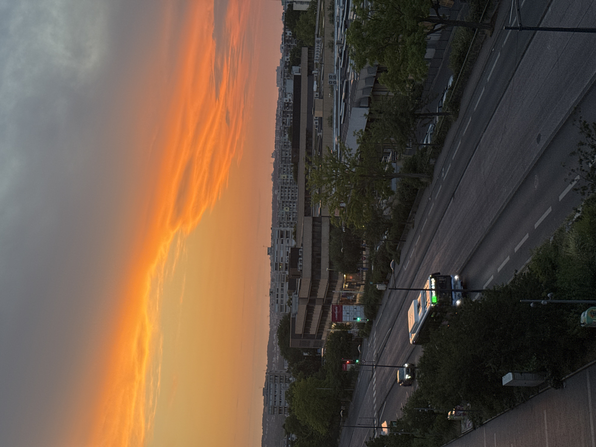 The image depicts a street scene in an urban environment during a sunset. The sky is dominated by vibrant orange and yellow clouds, transitioning to a gray, overcast sky. In the foreground, a paved road runs horizontally across the frame, with a sidewalk and green spaces to the left. Several trees line the right side of the road, obscuring some of the buildings behind them.
Vehicles are present on the road. A white bus with its headlights on is traveling on the right side of the road. A dark-colored car is seen ahead of the bus, also traveling in the same direction. Further down the road, other vehicles appear to be parked. Traffic lights are visible, with one showing green.
Behind the trees and the road, there are several multi-story buildings. Some of these buildings appear to be commercial or office spaces, with visible windows and architectural details. In the background, a skyline of apartment buildings stretches across the horizon, with some residential blocks visible on a gentle slope. The overall impression is of a suburban or edge-of-city setting. The image appears to have been taken from an elevated perspective, looking down onto the street and surrounding area.