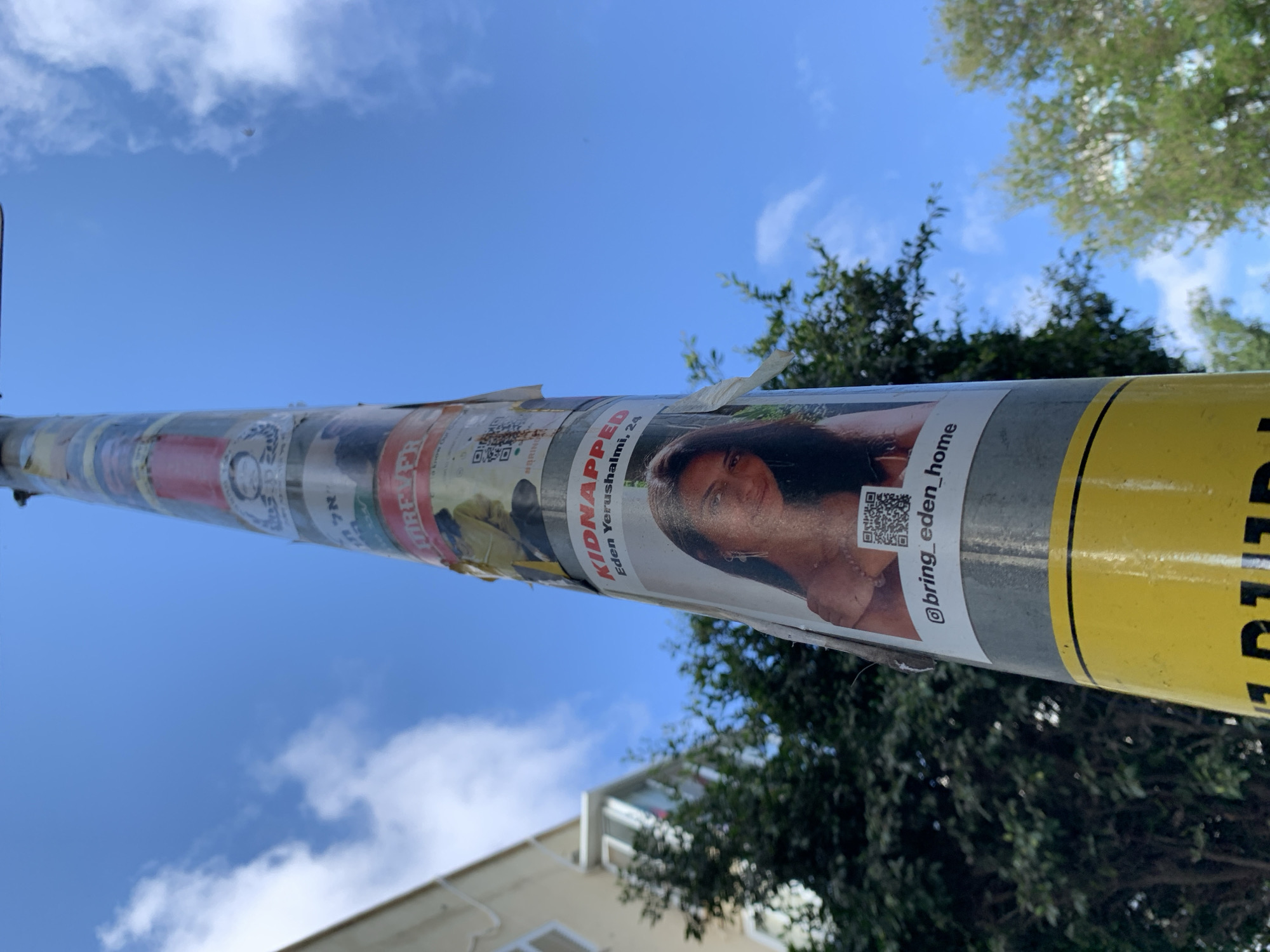 The image shows a utility pole covered in posters and stickers in Tel-Aviv, Israel. A prominent poster displays a photo of a young woman, identified as Eden Yerushalmi, age 24, with the word "KIDNAPPED" prominently displayed. The poster also includes a QR code and the Instagram handle "@bring_eden_home." The pole is situated outdoors, with blue sky and scattered clouds visible in the background, along with trees and parts of buildings.
