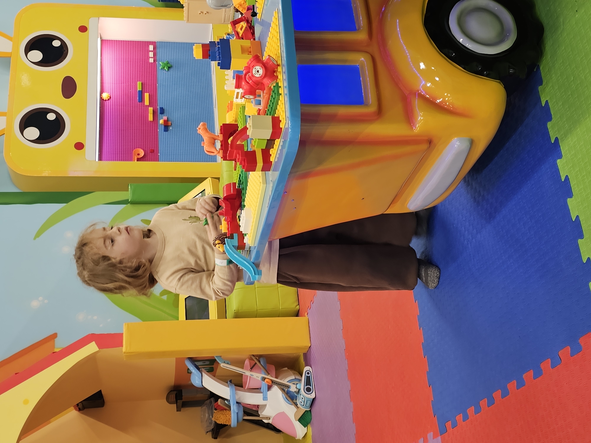 A young child with curly blond hair is standing at a play table in a brightly colored playroom. The child is wearing a tan long-sleeved shirt and brown pants. The table is shaped like a yellow car with a face on the front, featuring large, round eyes. On the table are various building blocks and toys. The playroom has interlocking foam mats in different colors on the floor (red, purple, blue, and green). The walls are painted light blue with green stylized grass designs. The lower part of the wall has additional play structures. The location is Khimki, Russia.