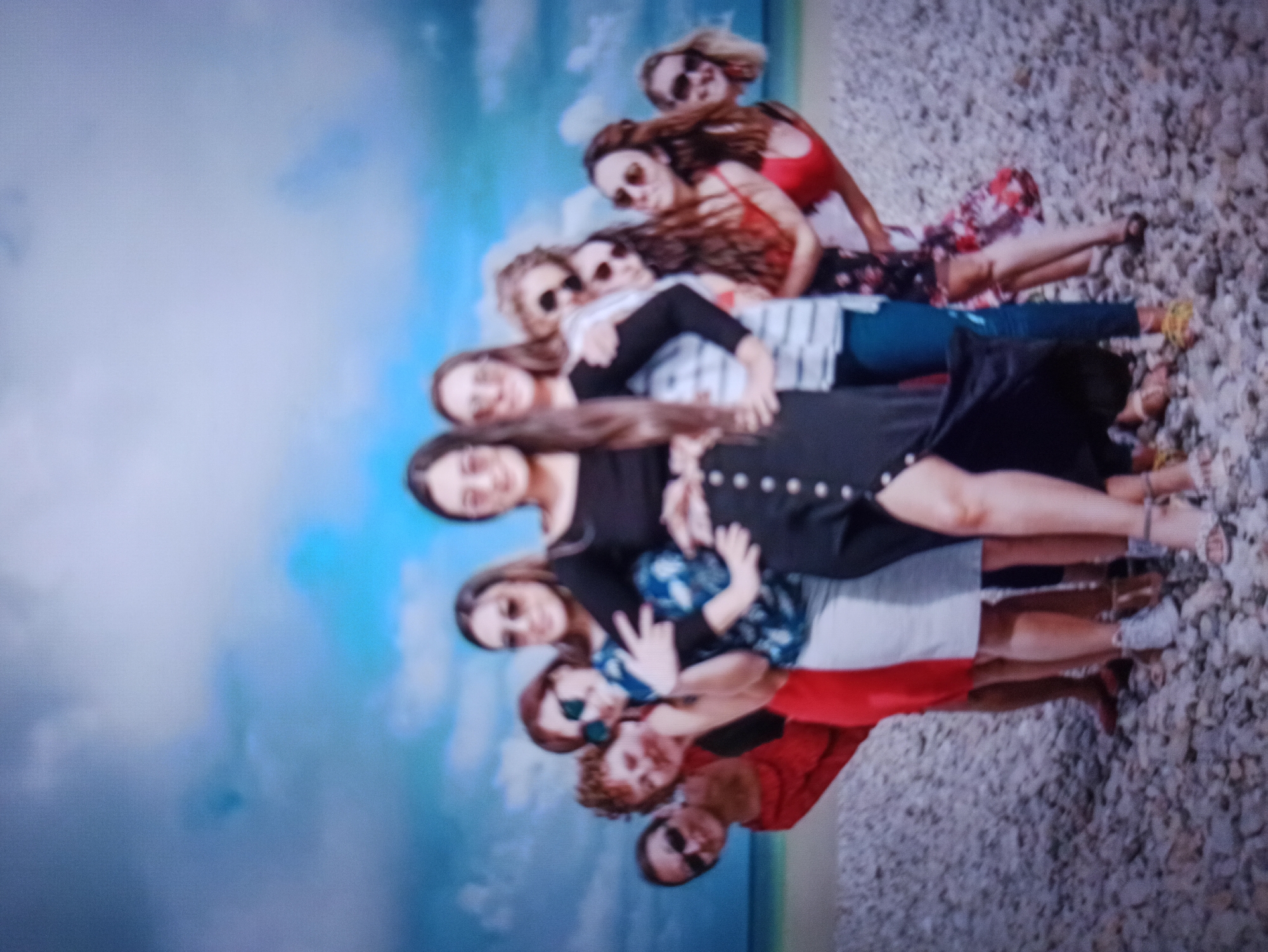 A group of ten women are posing for a photo on a rocky beach under a cloudy blue sky. The women are arranged in a semi-circle, with one woman in the center wearing a black off-the-shoulder top and a black skirt with white buttons. Several of the women are wearing sunglasses. The background shows a body of water and a cloudy sky. The image is slightly blurry.