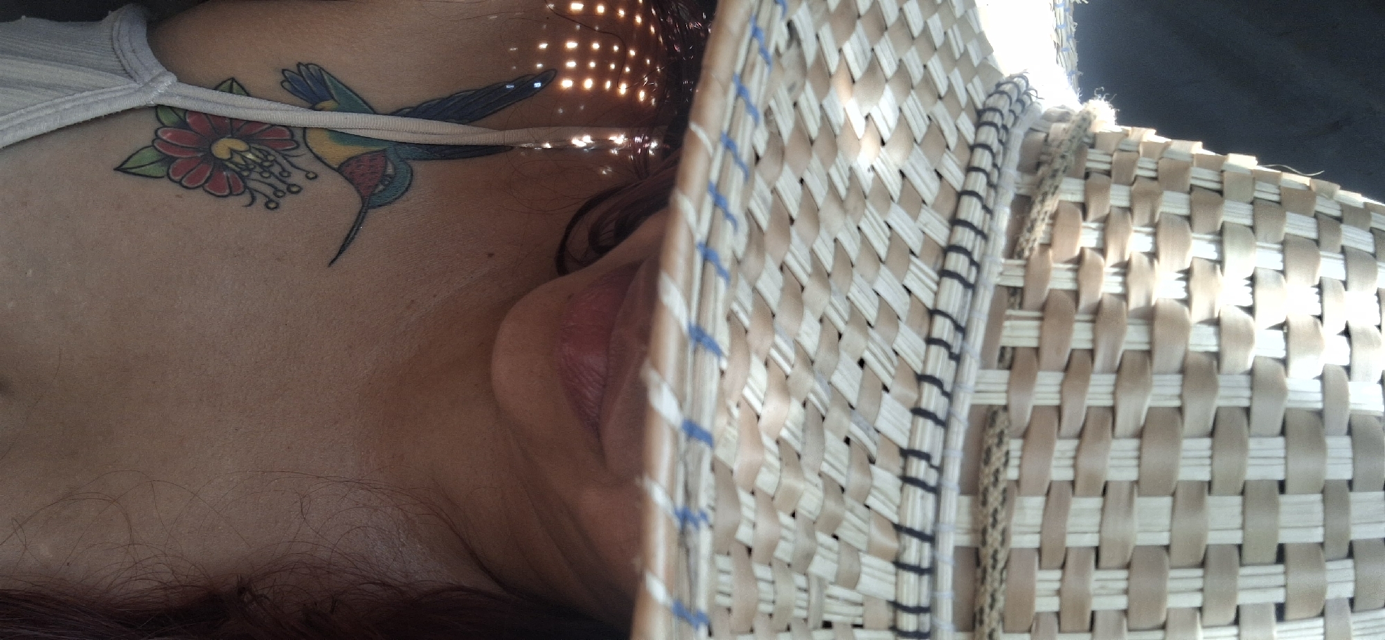 A person lies on their side with a woven basket held in front of their face. A hummingbird tattoo is visible on their chest. Light shines through the basket, creating spots on their face. The location is not discernible from the image.