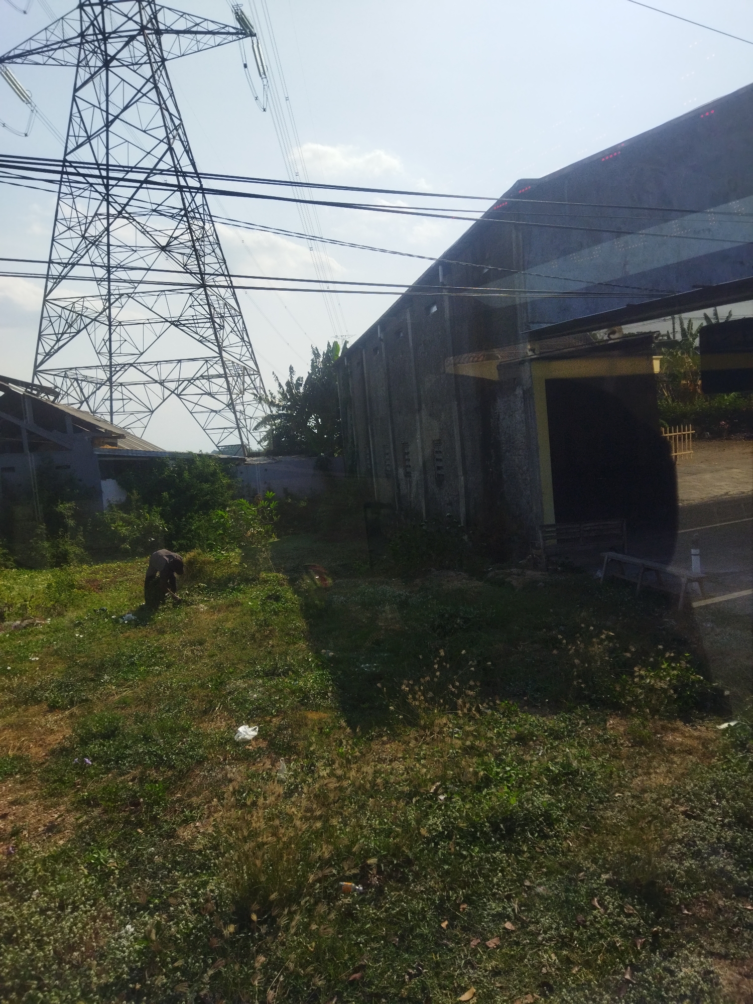 In the city of Kedungwinong, Indonesia, a person is seen working in an overgrown grassy area. The individual, appearing to be male and wearing dark clothing, is bent over, possibly tending to the vegetation or picking up debris. In the background, a massive electrical tower dominates the skyline, with numerous power lines stretching across the frame. Adjacent to the tower, a long, concrete building stands, its facade showing signs of age and weathering. The surrounding environment is characterized by lush, untamed greenery, with various plants and trees growing in abundance. Scattered white plastic bags are visible on the ground, suggesting litter or discarded materials. The scene is captured under a bright, clear sky, indicative of daytime.