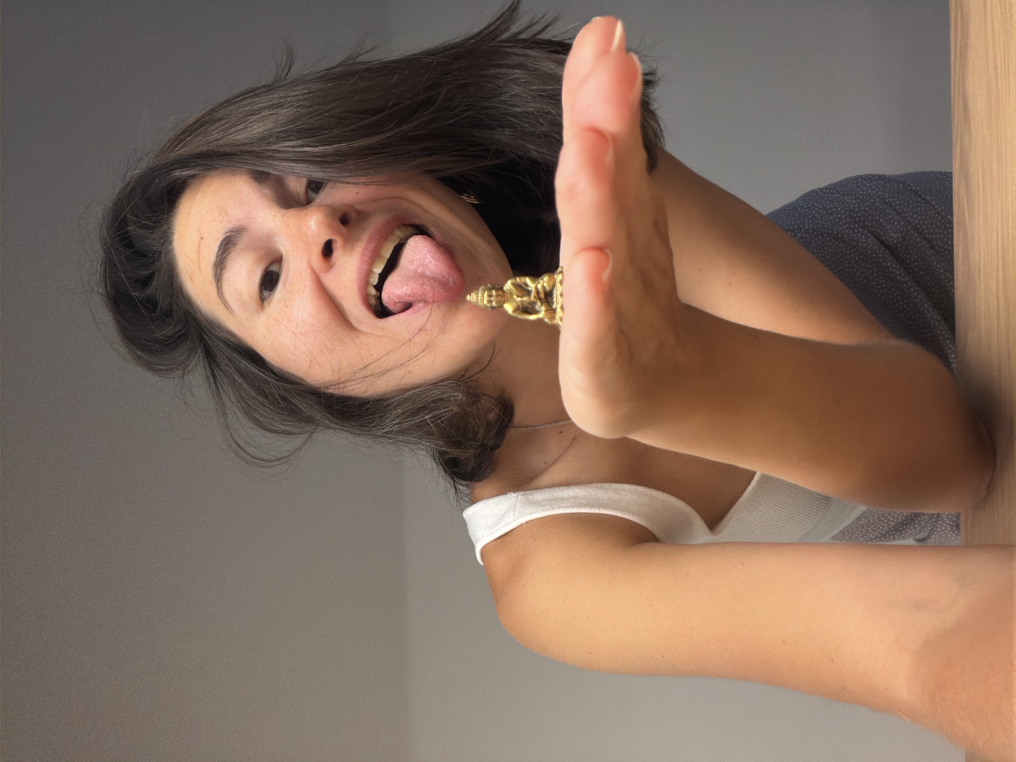 A woman with short, dark hair and fair skin is sticking her tongue out and holding a small, golden Buddha statue between her tongue and a raised hand. She is wearing a white tank top. The background consists of a light gray wall, a blue patterned surface, and a light wood-colored edge of what appears to be furniture. The photo is taken from a low angle, looking up at the woman. There is no indication of the location being Budva, Montenegro.