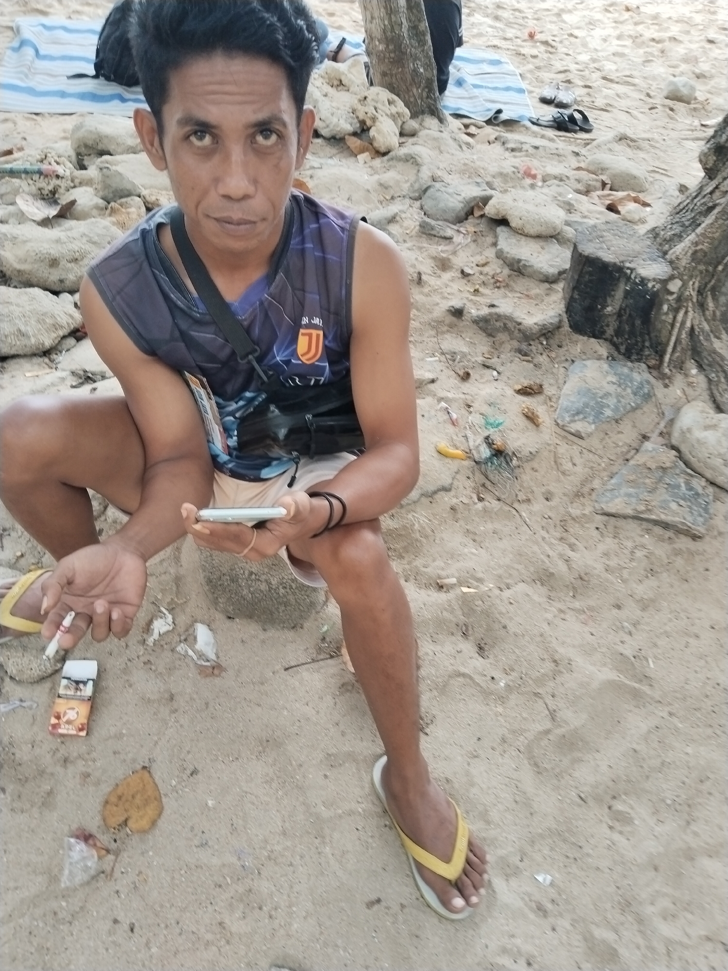 A man with dark hair is sitting on a sandy beach in Prigi, Indonesia. He is wearing a sleeveless shirt and shorts, and holding a cigarette and a mobile phone. A pack of cigarettes is lying on the sand next to him. In the background, there are rocks, trees, and a striped blanket. The man appears to be looking at the camera.