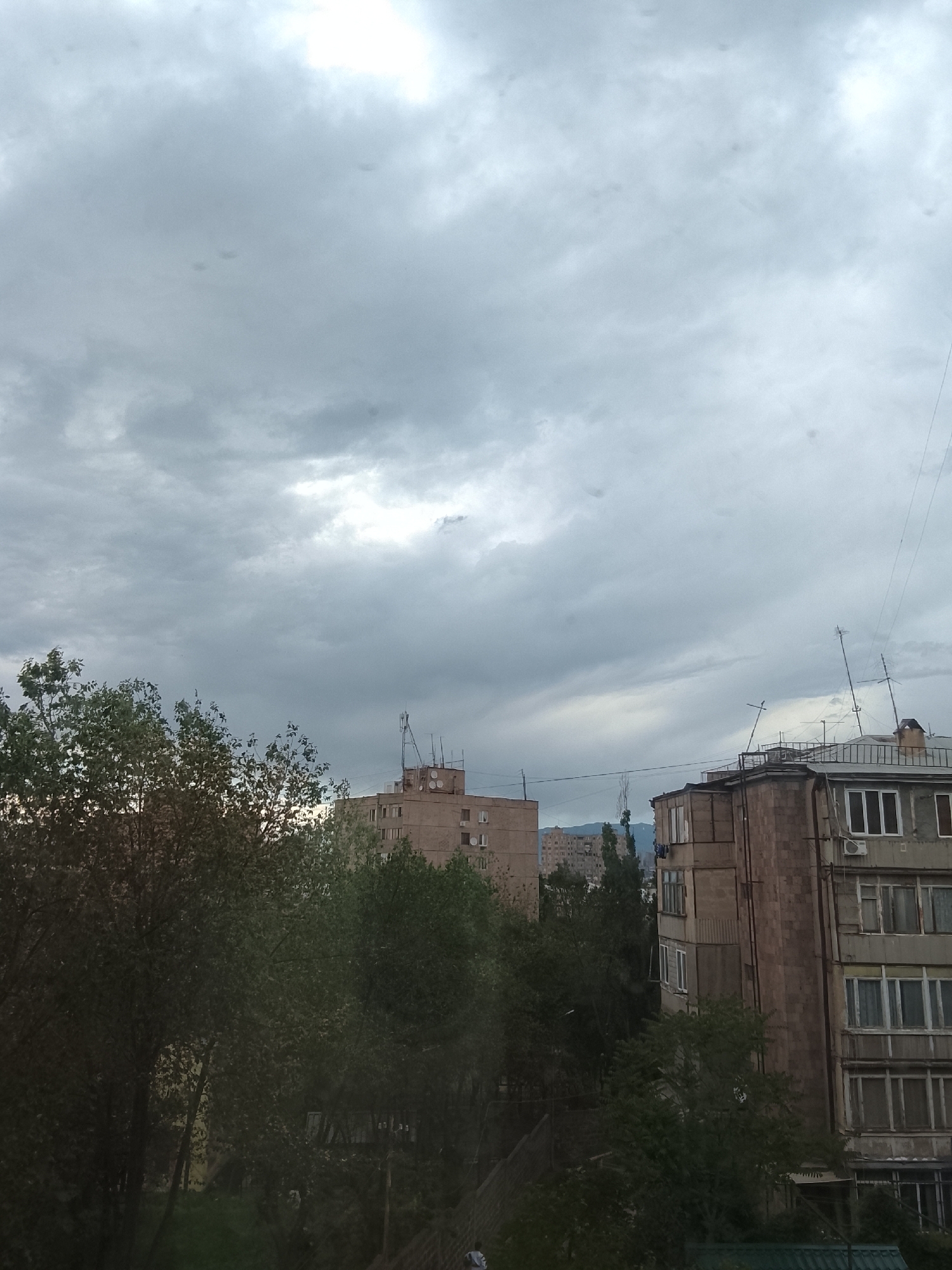 The image captures an overcast urban landscape during the daytime. The sky is dominated by thick, grey clouds with brighter patches, suggesting diffused sunlight. Several multi-story buildings are visible, partially obscured by lush, green trees and dense foliage that occupy the lower half of the frame.
In the mid-ground, a light-colored building features a flat roof with an array of communication antennas. To the right, another multi-story building, likely residential, showcases numerous windows, enclosed balconies, and air conditioning units. Its facade appears to be constructed from stone or concrete panels. Distant mountains or hills are visible on the horizon, partly veiled by the atmospheric haze. Thin utility wires or cables stretch across the sky on the right side of the image.
A small, indistinct human figure can be seen in the bottom left corner, partially hidden by trees, implying pedestrian presence. Several small, dark, irregular spots are visible against the cloudy sky, consistent with dust on the camera's lens or sensor. No legible text is visible within the image. The overall atmosphere is quiet and subdued, indicative of an overcast day in what appears to be an older, possibly post-Soviet urban environment.