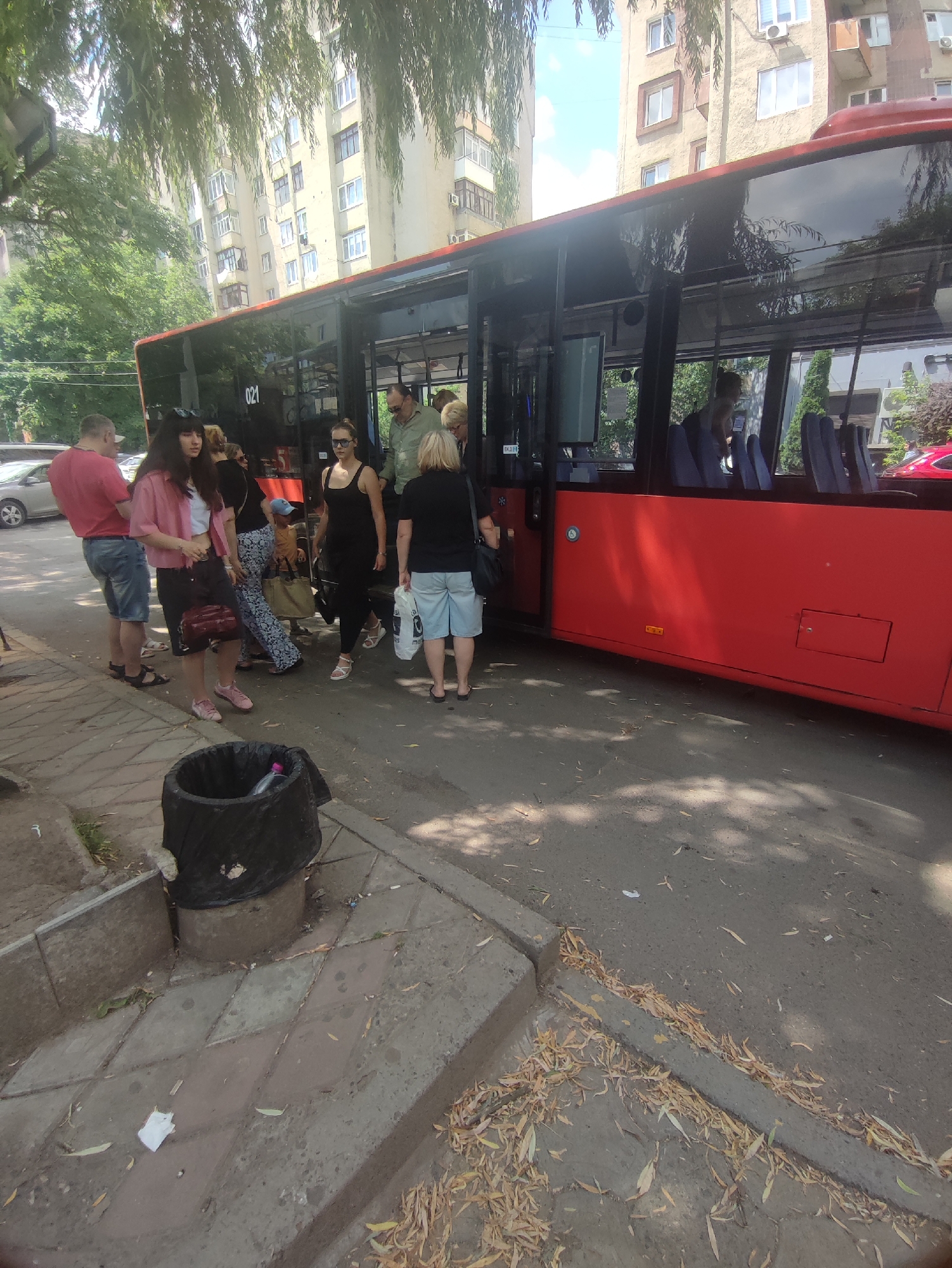 The image depicts a group of people boarding a red bus at a bus stop on a sunny day. The bus, identified by the number "021" on its side, is parked next to a sidewalk lined with trees. In the background, there are multi-story buildings characteristic of urban residential areas. A trash can is visible on the sidewalk. The people are dressed in casual summer clothing, with some holding bags. The scene suggests a typical urban scene in Chernivtsi, Ukraine, likely a bus stop on a street with residential buildings.