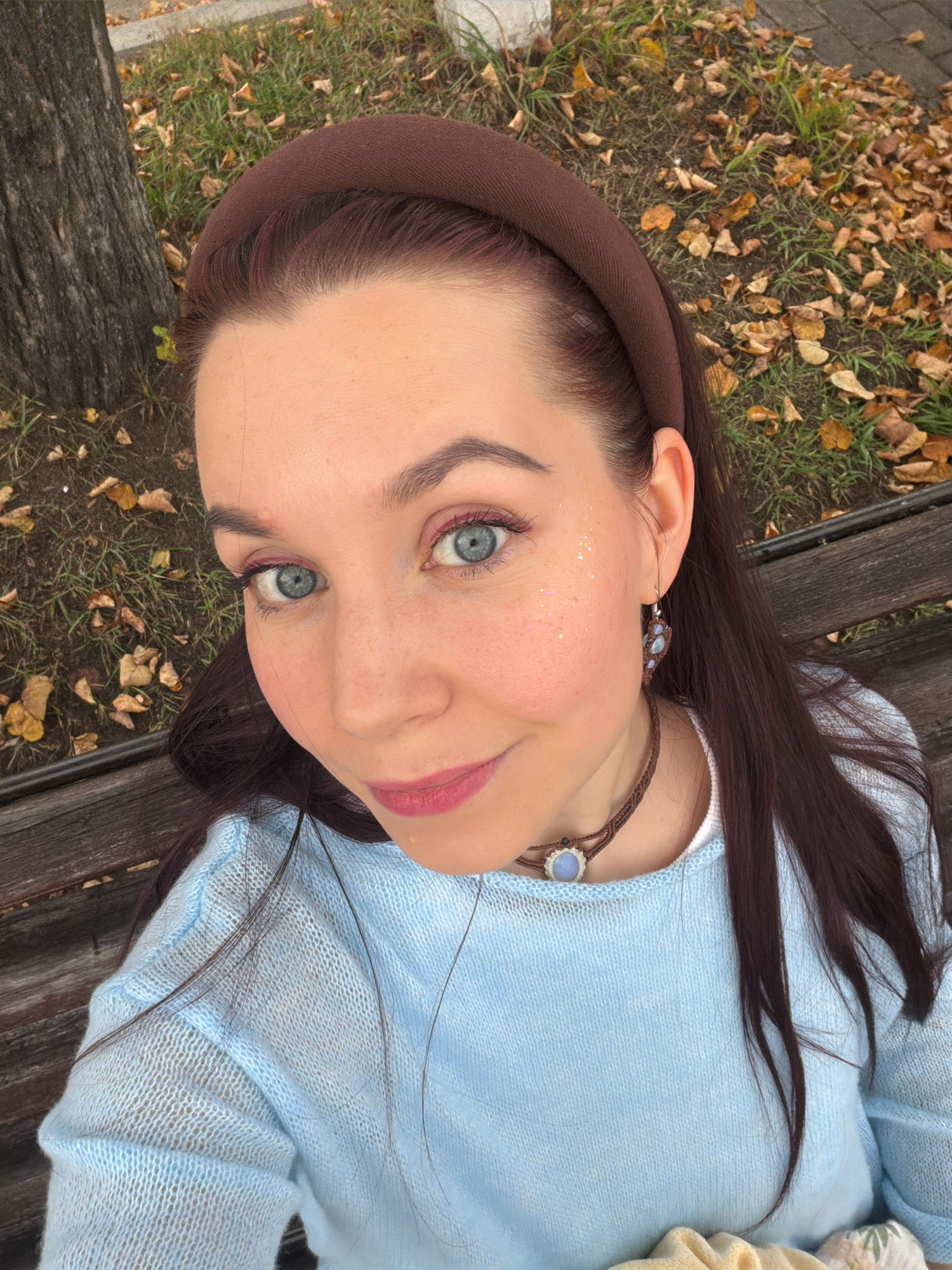 A young woman with light blue eyes, fair skin, and dark brown hair is taking a selfie outdoors on a wooden bench in Chaikovsky, Russia. She is wearing a light blue knitted sweater and a brown headband. She has pink eyeshadow and glitter on her cheeks. The background shows fallen autumn leaves and a tree trunk, suggesting the setting is a park or street. She is also wearing a macrame necklace with a light blue stone and a pair of dangle earrings with similar stones.