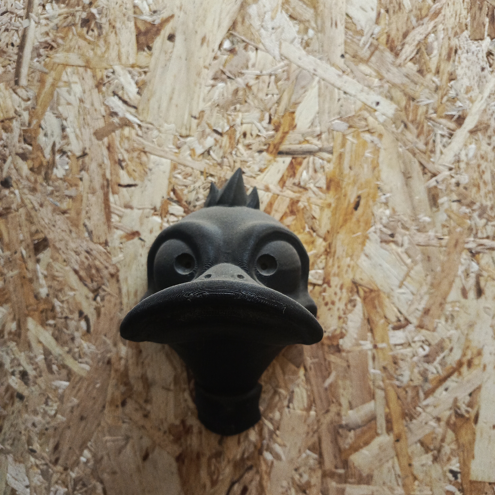 A black, 3D-printed cartoonish duck head is mounted on an oriented strand board (OSB) wall. The location is not discernible as being in the city of Chaikovsky, Russia based solely on this image. The duck head has large, round eyes with holes in the center, a wide, flat bill with horizontal ridges, and three small crests on top of its head. The OSB wall has a random pattern of wood chips and varying shades of tan and brown.