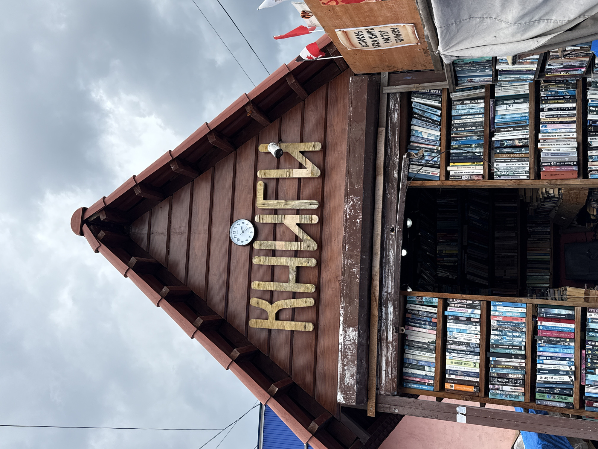The image shows a wooden stall filled with books. The stall has a gable roof and the word "КНИГИ" (Books) is written in large wooden letters across the front. A white clock is mounted above the letters, and a security camera is attached to the wall near the letters. Several shelves inside the stall are packed with books, some of which are also displayed outside in front of the stall. The stall is located outdoors, with a cloudy sky visible above. The text on a rolled scroll on the right side of the stall reads "ИСТОЧНИК ВСЕЙ КНИГИ РАСТИТ ЧЕЛОВЕКА" which translates to "The source of all books nurtures man." This is an outdoor bookstore in the city of Izhevsk, Russia.