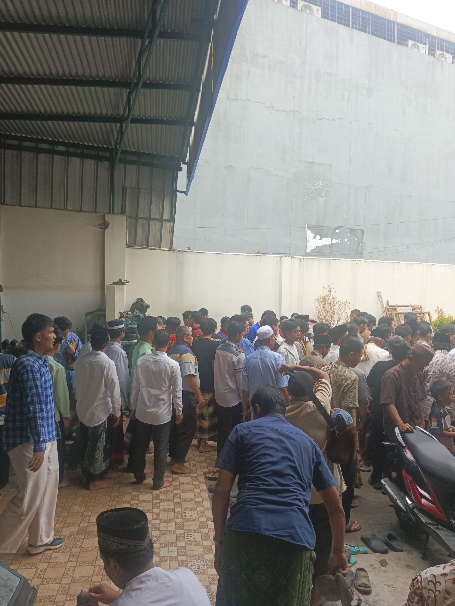 The image captures a gathering of people, predominantly men, in what appears to be an outdoor covered area adjacent to a building. The architecture suggests a location in South Tangerang, Indonesia, likely a mosque or a communal gathering space.
In the foreground, a man wearing a blue plaid shirt and cream-colored pants is walking from left to right. To his left, several individuals are standing and talking. Directly behind him, a group of men are clustered together, some wearing white shirts and peci hats, typical of religious attire.
The environment features a corrugated metal roof supported by green metal beams, casting shadows. The floor is tiled with a patterned design. Behind the people, a pale beige wall of the building is visible, with some cracks and patches on its surface. Further in the background, a portion of a large, pale gray building with visible air conditioning units can be seen.
Several pairs of sandals and shoes are scattered on the tiled floor, indicating that people may have removed them upon entering a prayer area or for comfort. A motorcycle is parked on the right side of the frame. The overall atmosphere suggests a social or religious event, possibly a gathering after a prayer service or a community meeting. The lighting is overcast, creating a soft illumination.