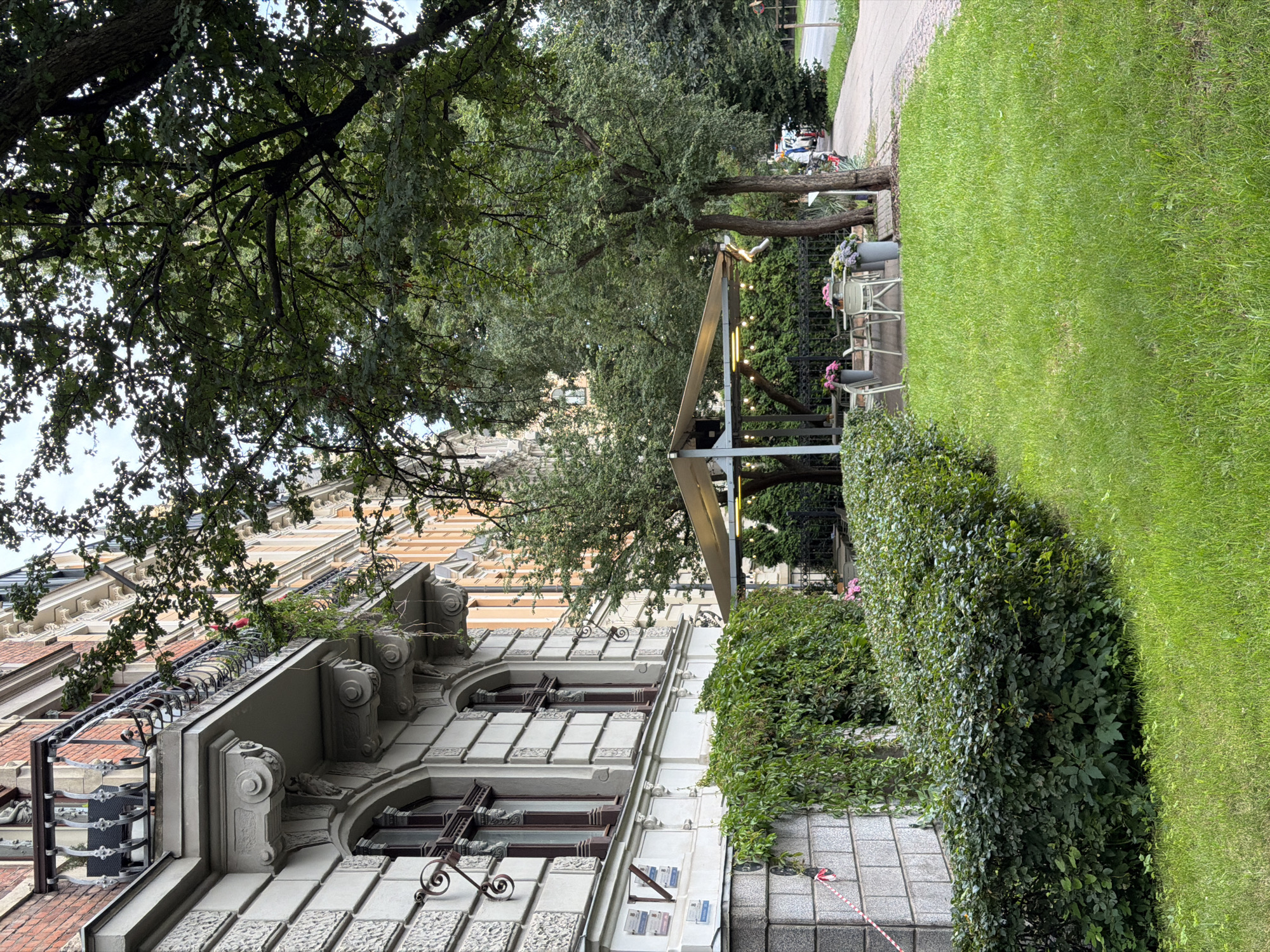 This is a medium-shot, eye-level photograph capturing an outdoor cafe seating area adjacent to a building and a tree-lined street. The building on the left is a multi-story structure with ornate architectural details, including a balcony with a wrought-iron railing and decorative carvings. The lower portion of the building is covered in ivy.
In the center of the image, an outdoor cafe is set up under a light-colored awning. It features several white tables and chairs, some with potted plants. String lights are suspended above the seating area.
To the right of the cafe, a paved walkway runs parallel to the street. Trees with dense green foliage line the walkway and the area around the cafe. Beyond the walkway, a street is visible with some vehicles parked or in motion, including what appears to be a motorcycle and a car. The environment suggests an urban setting, and the architectural style of the building is consistent with the Old Town or nearby historic districts of Riga, Latvia. The overall scene depicts a pleasant, possibly late afternoon or early evening setting in a city.