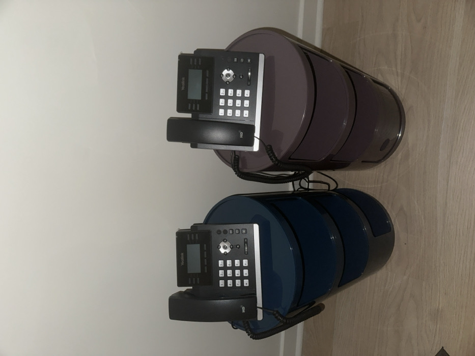 Two Yealink office phones sit atop cylindrical storage units, one navy blue and the other a mauve-grey color. The units have a circular top and bottom with a slightly convex side, separated by a band. The phones are black and have a large keypad and handset. They are positioned near a white wall and light wood floor.