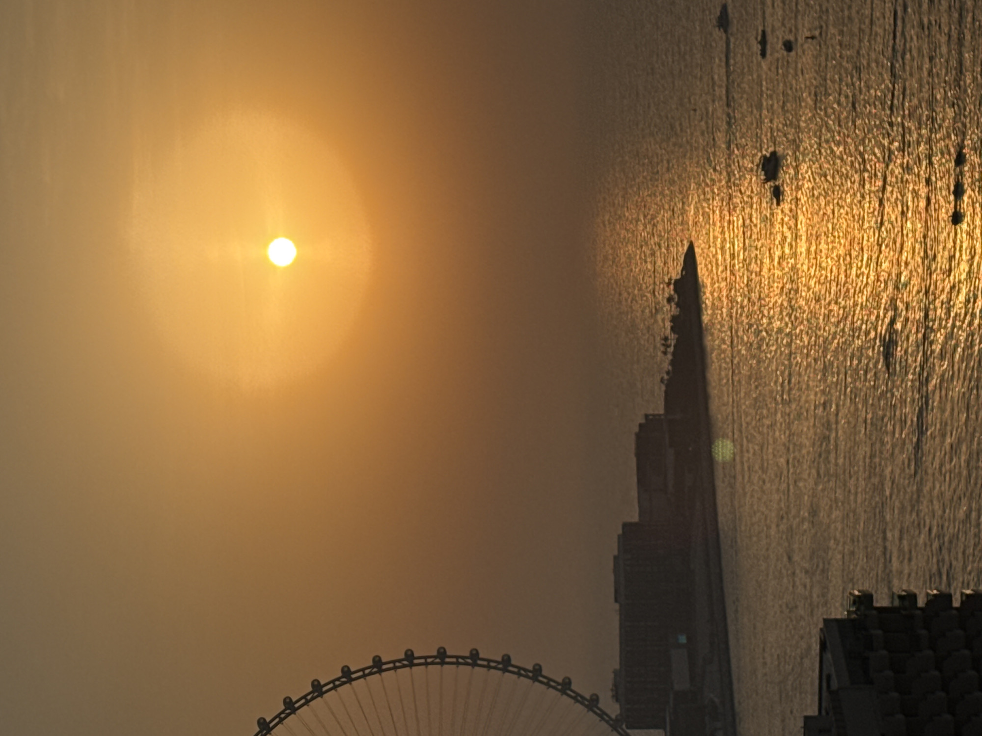 This is a vertical, eye-level, medium shot of a sunset over the sea in Dubai, United Arab Emirates. The sun is a bright, hazy orb in the upper right quadrant of the frame, casting a golden glow across the scene and reflecting off the water.
In the left side of the frame, the lower portion of a large Ferris wheel is visible, its spokes and cabins silhouetted against the warm sky. Along the distant shoreline, a row of dark, multi-story buildings is visible, with palm trees dotting the landscape in front of them. The buildings appear to be hotels or residential complexes.
The sea is relatively calm, with a shimmering reflection of the sunset creating a path of light across its surface. Several small boats are scattered on the water, some of which appear to be docked. In the foreground, a portion of a building with a dark, textured facade is visible on the left.
The overall impression is one of tranquility and the vibrant colors of a Dubai sunset.