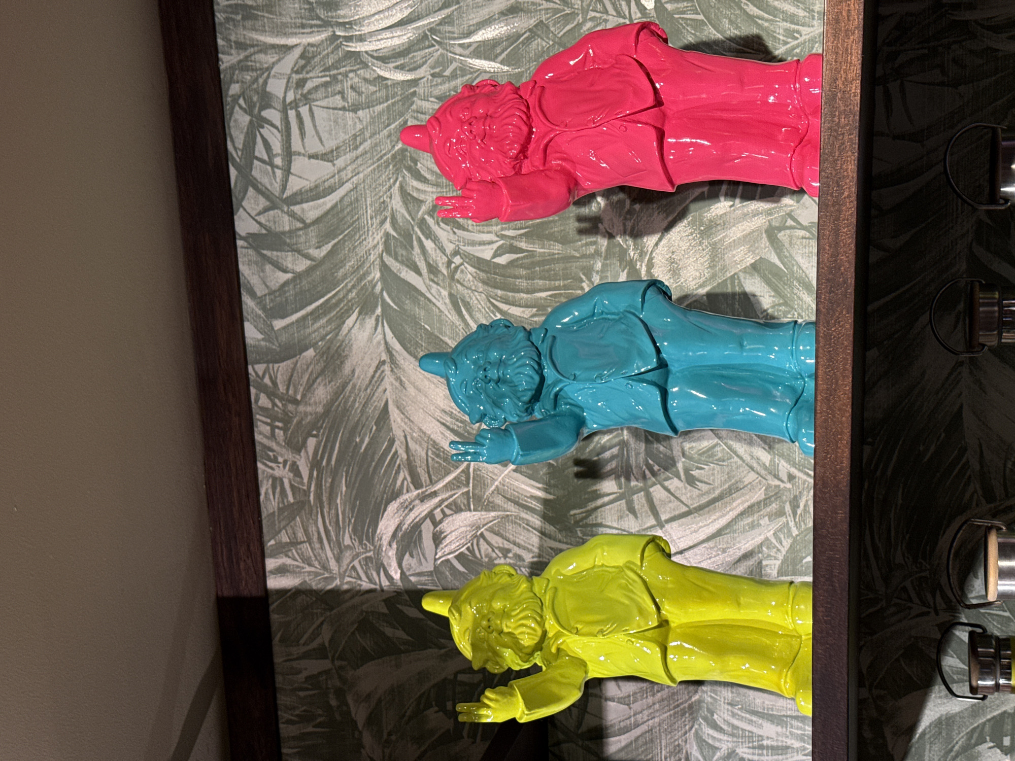 The image shows three painted gnome figurines aligned vertically in front of a wall with a floral pattern. The gnomes are colored pink, turquoise, and yellow, respectively. The composition is framed by a dark brown border on the left and the side of a dark wooden structure with metallic accents on the right. The image appears to be taken indoors, but the exact location in Kuta, Indonesia cannot be determined from the visual information.