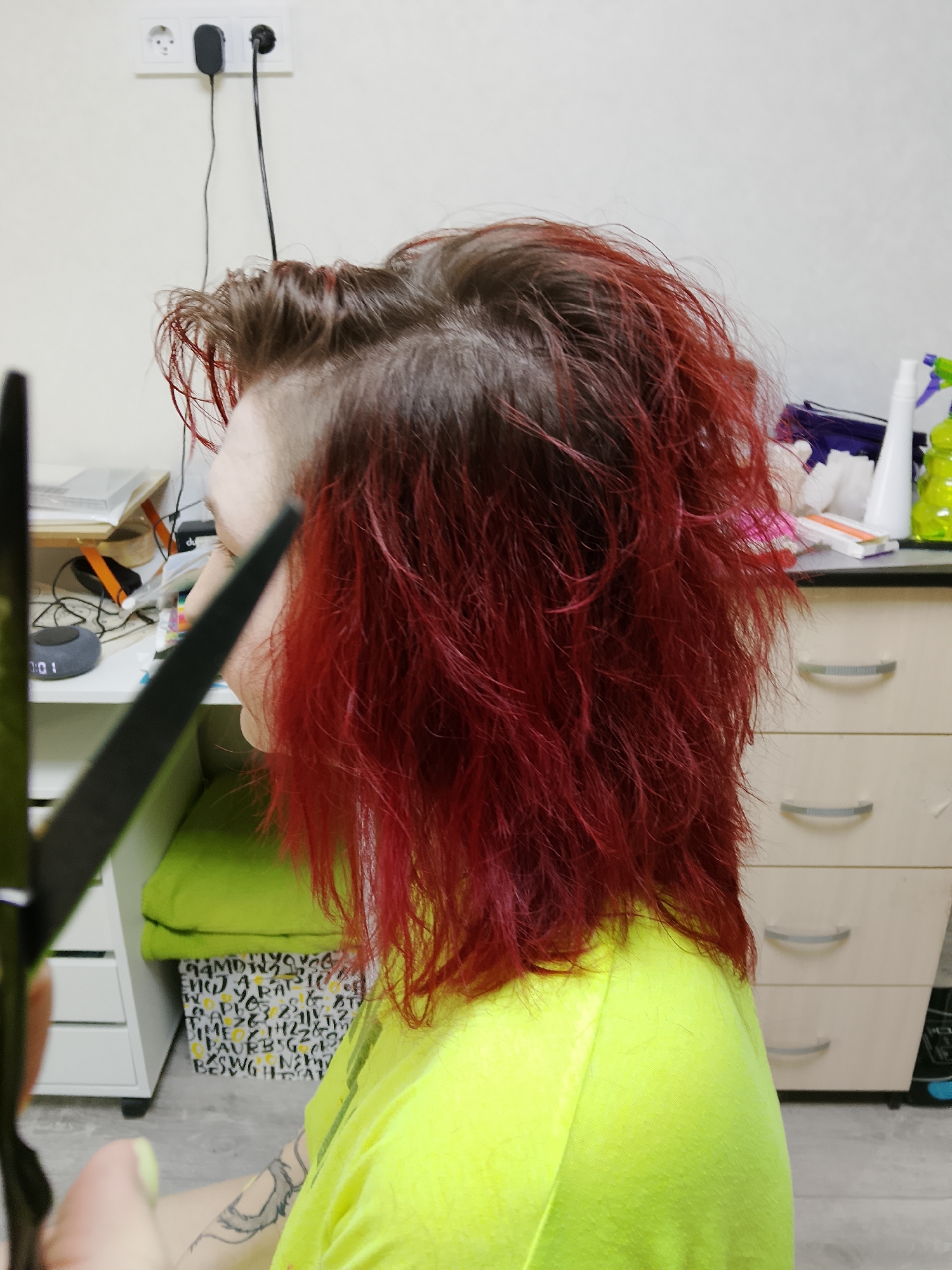 A person with red hair and a shaved side is sitting in a room. The person is wearing a yellow-green shirt. A pair of scissors is held up to their hair. The room contains a desk with a clock and other items on it, a white cabinet, a green cushion, a spray bottle, and other objects. The wall has an electrical outlet with plugs. The location is Chaikovsky, Russia.