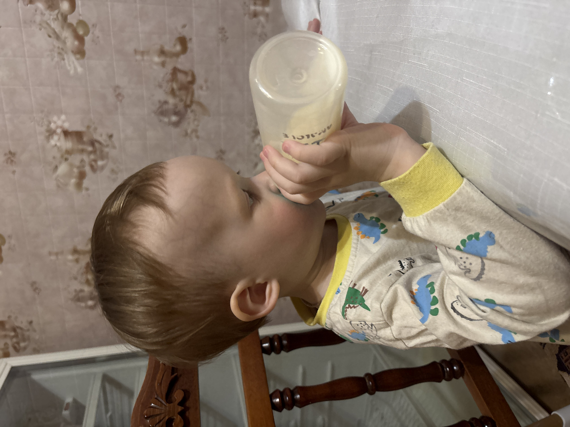 An image, rotated 90 degrees clockwise, depicts a young child, likely a toddler, drinking from a baby bottle. The child has light brown hair and is wearing pajamas featuring blue and green dinosaur patterns and yellow cuffs. A hand is seen holding the transparent bottle, which contains a light yellow liquid, presumably milk or formula. Visible text on the bottle reads "PROJECT" and partially "AM_C_LE". The child's ear is visible, and their focus is entirely on the bottle.
The background features patterned wallpaper with faint, abstract floral or organic designs in muted brown tones. Below the child, a dark wooden chair or furniture piece with turned legs and carved details is partially visible. To the right, a white, subtly textured fabric, possibly a curtain or bedsheet, fills a significant portion of the frame. The scene is set indoors, suggesting a domestic environment in Ферма, Russia, likely during the evening given the child's sleepwear. The lighting is soft and ambient.