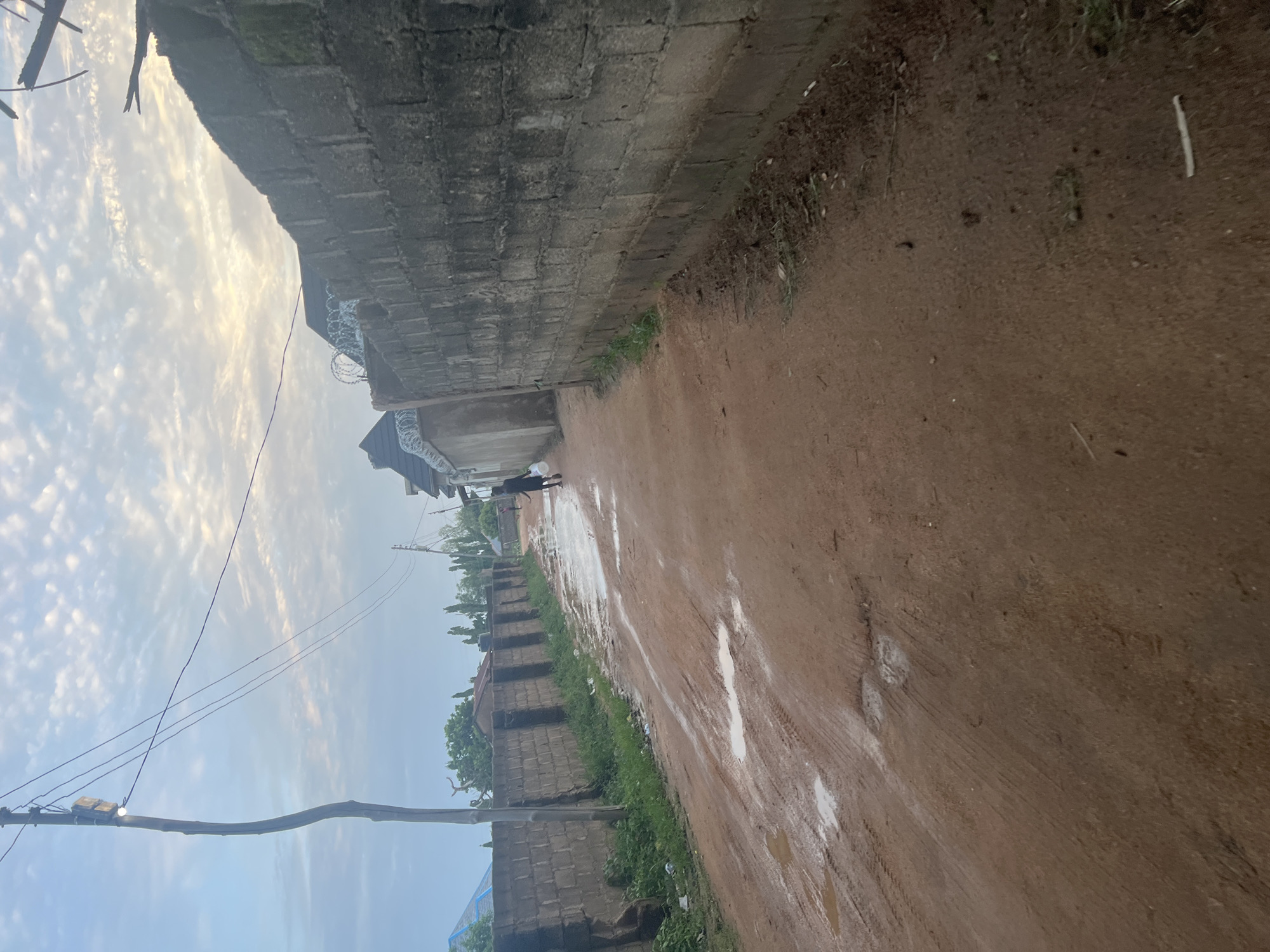 This image depicts a dirt road in Howayi, Nigeria, after a rainfall. The road is muddy and has several puddles. On the right side of the road is a tall, rough concrete wall. On the left, there is a lower brick wall and some vegetation. In the distance, a person is walking away from the camera down the road, carrying a white object in their right hand. A wooden utility pole with wires and a light fixture stands on the left side of the road. The sky above is cloudy with patches of sunlight.