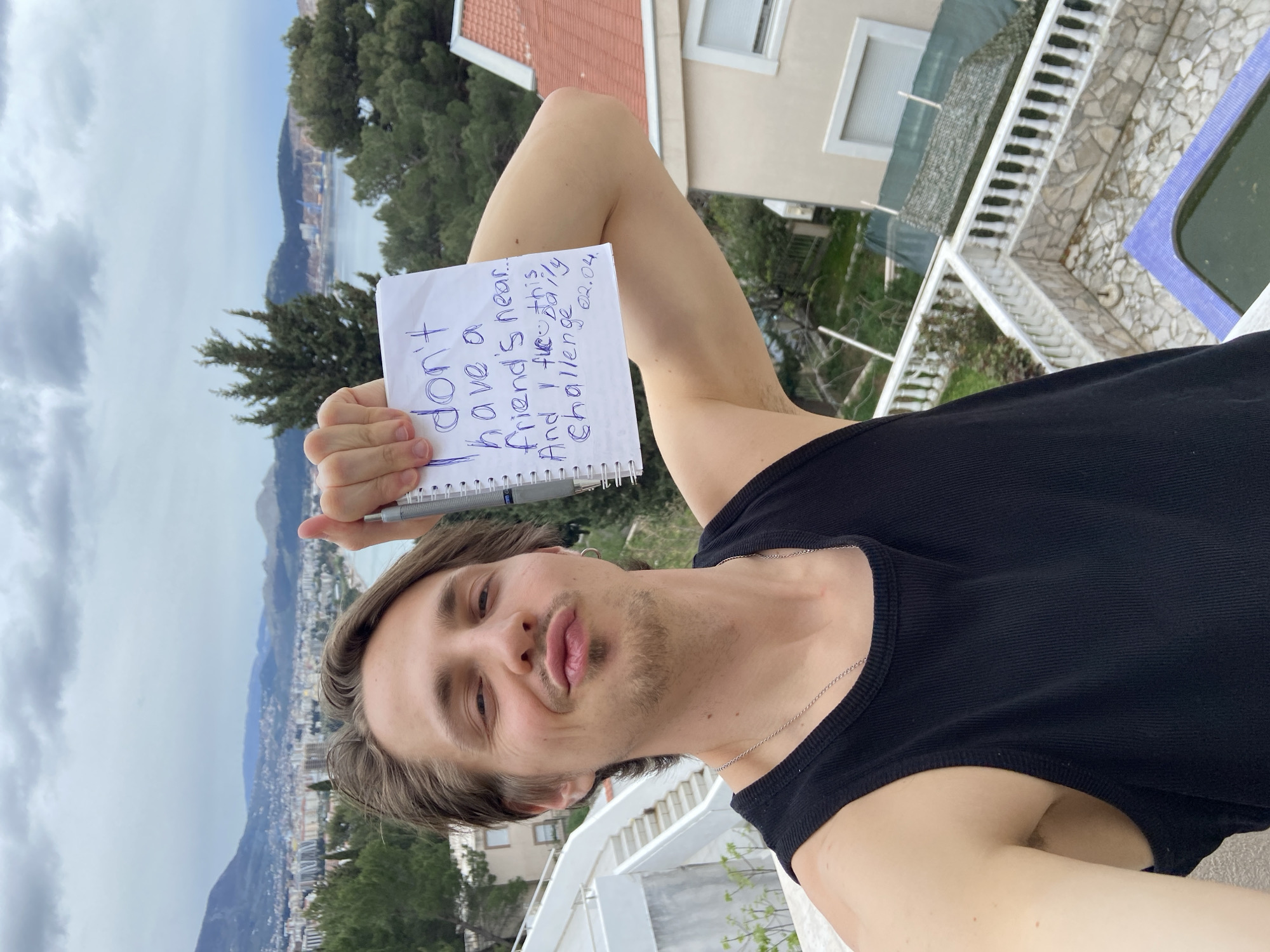 A man holds a spiral notebook with a handwritten message in a selfie. The message reads, "I don't have a friend's near. And I feel this... Daily challenge 02.04." He is wearing a black tank top and a silver necklace. He has brown hair, a slight beard, and is making a pouting face. Behind him is a view of a city by the water, with mountains in the background. The location appears to be on a balcony or rooftop with a white railing. A swimming pool is partially visible to the right. The city view includes buildings and the coastline. The location is Šušanj, Montenegro, as indicated by the city view.