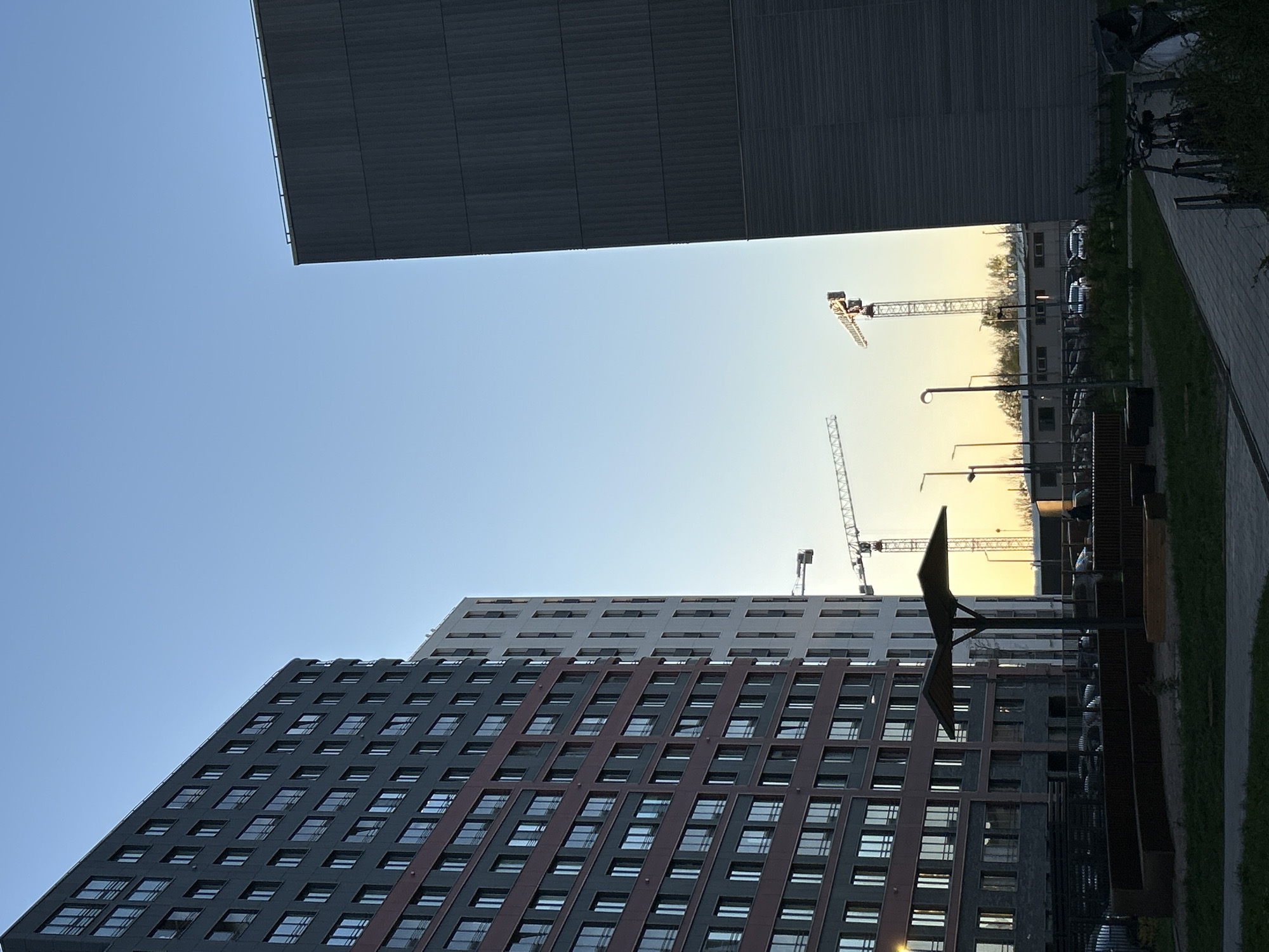 The image shows an urban view, likely in Saint Petersburg, Russia, based on the architectural style. A multi-story building with a dark facade and rows of windows dominates the lower portion of the frame, partially obscured by a modern, sculptural element in the foreground. Above, the sky transitions from blue to a pale yellow, suggesting either sunrise or sunset. Several construction cranes are visible in the distance. Another large building with a simpler facade can be seen in the upper portion of the frame, giving the image a sense of enclosure. A paved walkway and greenery are visible on the right side, indicating a pedestrian area. The scene appears to be a combination of residential and construction zones.