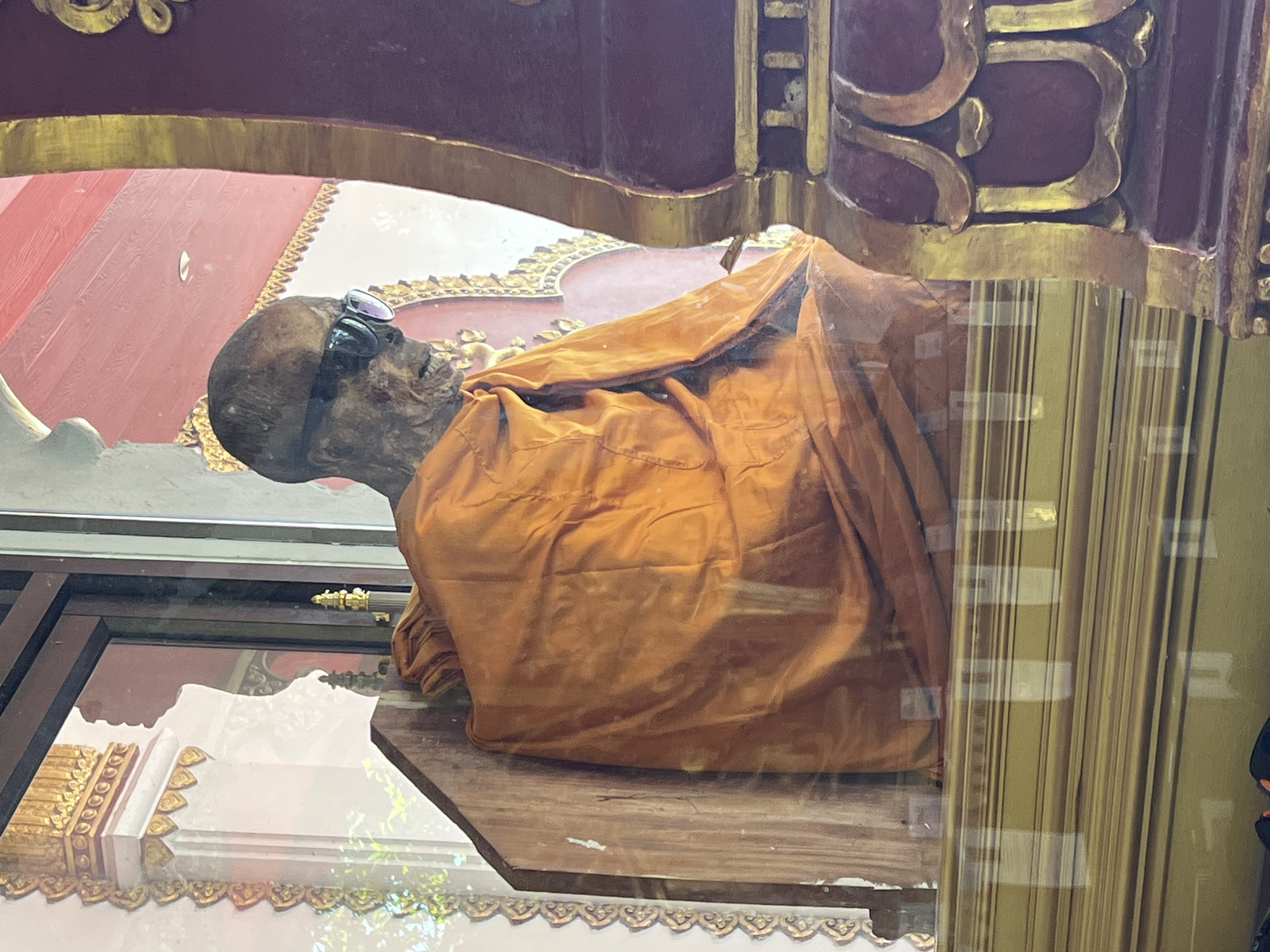 The image shows a mummified Buddhist monk seated in an orange robe and wearing sunglasses. The monk is displayed inside a glass case, possibly a temple or museum, in Baan Hua Thanon, Thailand. The background features ornate golden decorations and red architectural elements, suggesting a place of cultural or religious significance. The monk's body is visibly preserved, with dried skin and exposed bones. The overall scene is presented in a neutral, documentary style.