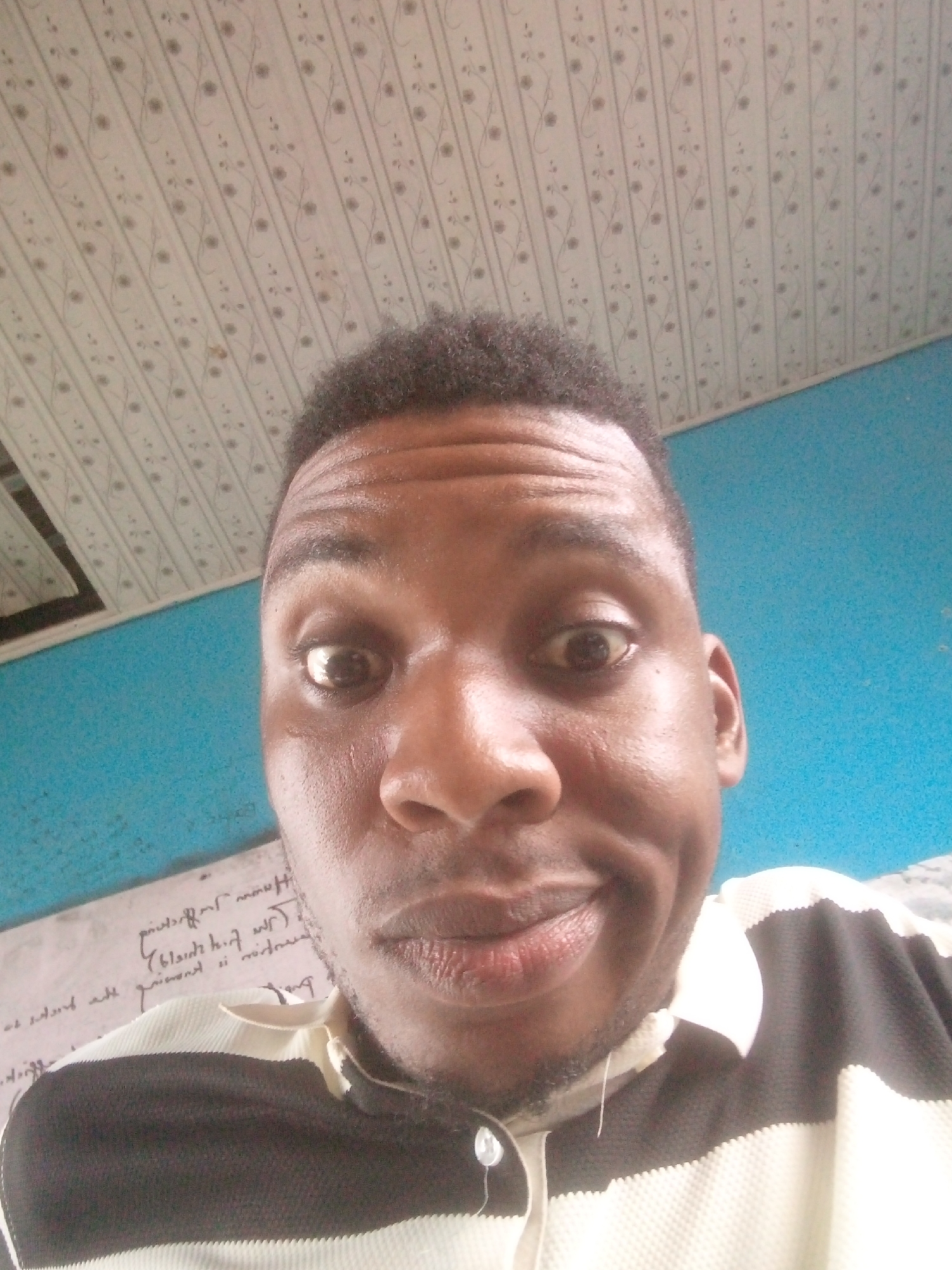 A close-up selfie photograph shows a dark-skinned man with short, dark hair. He has brown eyes that are wide open, a broad nose, and a slight smile. He is wearing a black and white striped shirt with a visible button. The background includes a white ceiling with a repetitive floral pattern, a teal-colored wall, and a white surface with illegible writing on it. The location appears to be indoors.