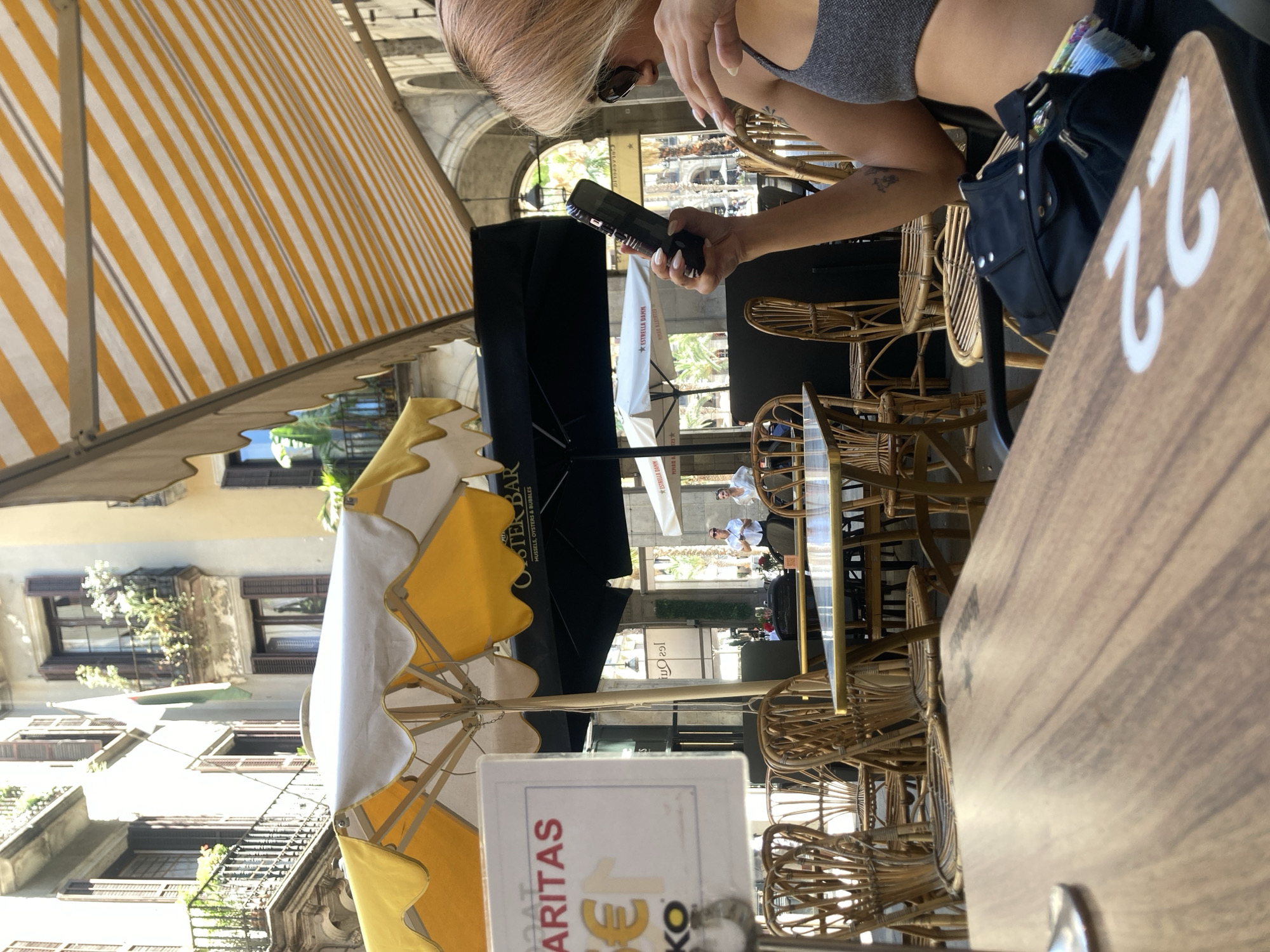 This media file shows an outdoor seating area of a restaurant or bar in Barcelona, Spain. A person with blonde hair, wearing sunglasses and a grey top, is seated and holding a black electronic device. The scene is partially covered by a yellow and white striped awning and a white and yellow umbrella. In the background, there are buildings with balconies and windows, suggesting an urban setting. Several wicker chairs are visible around tables, indicating a dining or lounging environment. A black bag is placed on one of the chairs. A table in the foreground has a sign with the number "22" on it. Visible text on signs includes "OSTER BAR", "MUSSELS, OYSTERS & BUBBLES", and "ARITAS €1 KO". The bright sunlight suggests it is daytime. The overall impression is a relaxed atmosphere at an outdoor establishment.