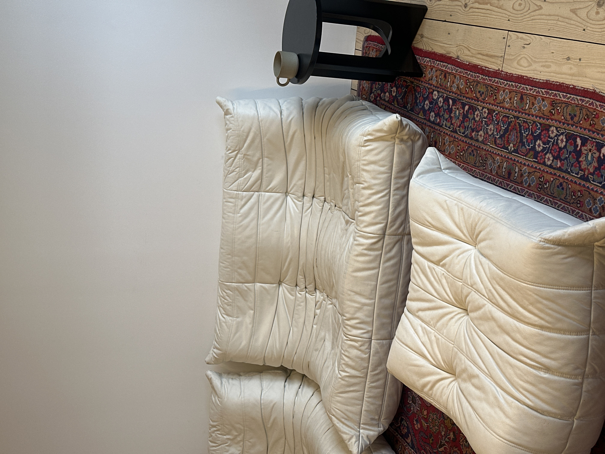 The image captures an interior living space, likely in London, United Kingdom, characterized by modern and comfortable furnishings. Two plush, off-white quilted sofas or armchairs, which appear to be Ligne Roset Togo style, dominate the foreground and midground. Their soft, creased texture suggests a comfortable, inviting design.
The furniture rests upon a richly patterned rug with intricate motifs in shades of red, blue, and brown, partially covering light-colored wooden floorboards. In the background, a black, angular side table holds a light grey ceramic mug. A plain white wall serves as a minimalist backdrop for the scene.
The lighting is soft and even, typical of an indoor daytime setting, without any harsh shadows. No people are present, and there is no specific activity taking place. The overall impression is one of a stylish, relaxed, and comfortable domestic environment. No visible text is present in the image.