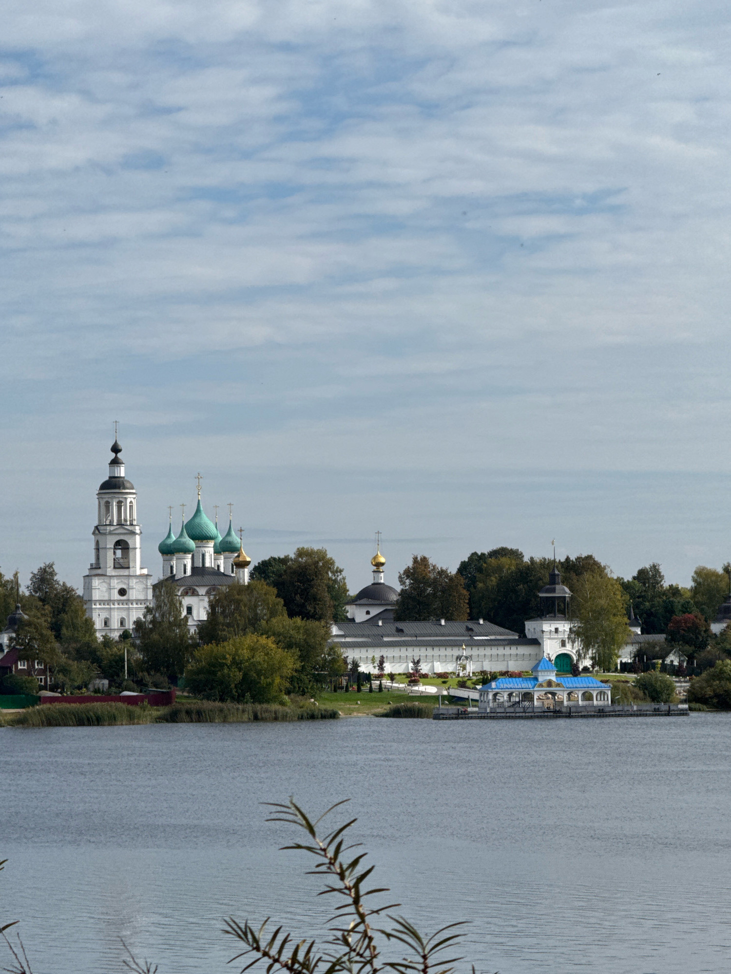 The image shows a view of the Uspensky Monastery in the city of Zhukovo, Russia, from across a body of water. The monastery complex features several white buildings with traditional Russian Orthodox church architecture, including a prominent bell tower with a black spire and multiple domes, some of which are turquoise. A brightly colored blue-roofed building, possibly a chapel or pavilion, is situated on the edge of the water, extending out over the lake. The monastery is surrounded by lush green trees, with some autumn foliage visible. The sky is cloudy, with streaks of blue peeking through. The foreground is dominated by the rippling surface of the water and a few branches of a plant in the lower right corner. There are no people visible in the scene.