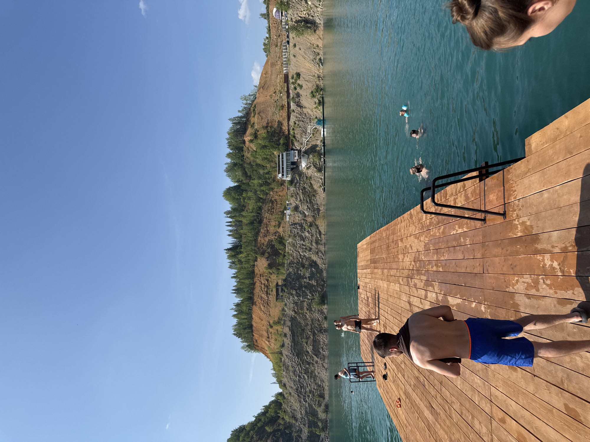 The image depicts a quarry lake in Karyer Izvestnyak, Russia, on a sunny day with a clear blue sky. In the foreground, a man wearing blue swim trunks and a towel around his neck stands on a wooden pier. Several other people are visible on the pier and in the turquoise water. Some are swimming, while others appear to be standing or walking on the wooden planks. In the background, a steep, rocky embankment covered with trees rises from the water. A building complex with white walls and a dark roof is situated on the embankment, along with a staircase leading down to the water. The overall scene captures a recreational day at a quarry.
