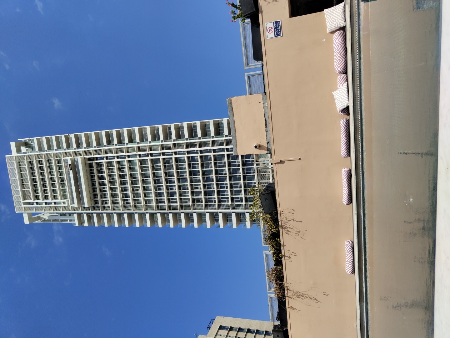 The image, captured in Tel-Aviv, Israel, depicts a modern urban scene looking upwards towards a tall skyscraper against a clear blue sky. The prominent, high-rise building on the left side features a contemporary design with a white facade, characterized by numerous floors of balconies and large windows arranged in a distinct grid pattern.
In the foreground, on the right, is a lower, tan-colored structure, likely a terrace or rooftop lounge. This area is equipped with several patterned cushions – a mix of rectangular pillows and cylindrical bolsters – placed along its edge, suggesting outdoor seating. The cushions display a light-colored geometric pattern. Two black box speakers are mounted on this wall, alongside a small security camera and some sparse greenery.
A "No Smoking" sign is prominently displayed on the tan wall. The sign features the universal no-smoking symbol and Hebrew text that reads "אין לעשן" (No Smoking) and "מחוץ למבנה" (outside the building).
The sky is a vibrant, cloudless blue, indicating bright and sunny weather during daylight hours. The scene is static, without any visible people or specific activity, focusing instead on the architectural elements and the serene urban setting. The overall impression is one of modern city living in clear, pleasant conditions.