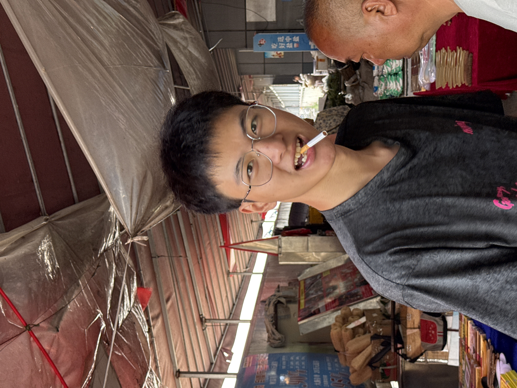 A young man with short black hair and glasses is in the foreground, biting down on a lit cigarette. He is wearing a dark gray t-shirt with a pink graphic design on the left side. To his right, partially visible, is an older man's head and shoulder, looking towards the young man. They appear to be in an outdoor market setting under a red and white striped awning. In the background, there are stalls with various goods, including what appear to be bags of produce, boxes, and signage with Chinese characters. A red triangular sign is visible in the mid-ground. The overall environment suggests a busy marketplace in Jiangjin, China.