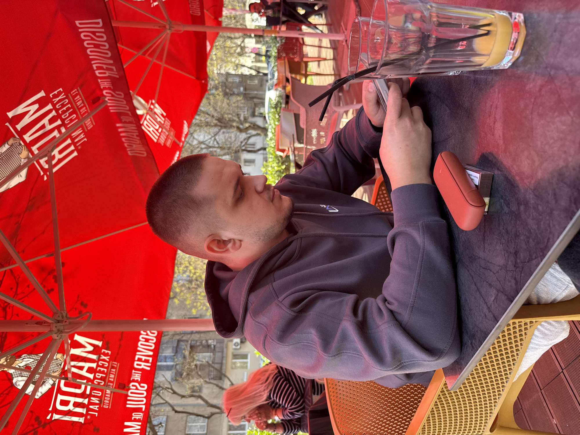 A man with a buzz haircut, wearing a gray hooded sweatshirt, is sitting at an outdoor cafe table. He is holding a mobile phone in his hands, his attention directed towards the device. On the table, there is a tall glass with a yellow liquid and two black straws, along with a small orange device and a pack of cigarettes next to it. A red umbrella with white writing is directly above him. In the background, there are buildings, trees, and other people, including a woman with blonde hair wearing a striped shirt. The café appears to be in an urban setting. The umbrella text suggests the café is branded with "WHAM!" possibly indicating a specific establishment. The location is likely in Plovdiv, Bulgaria.