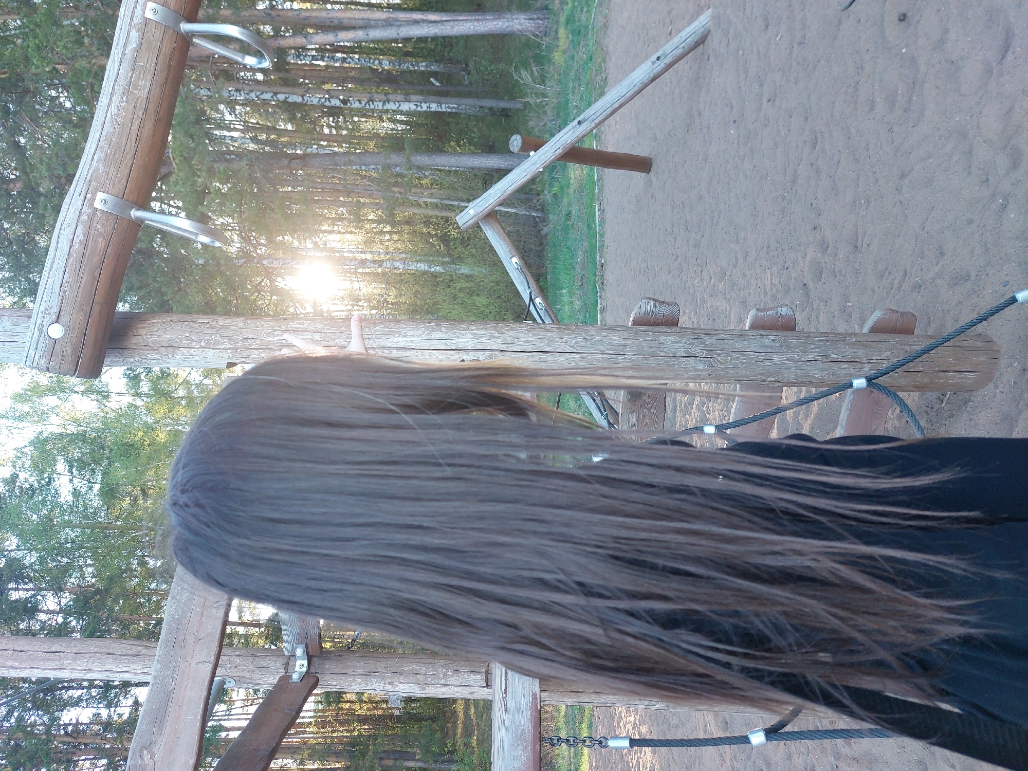 The image shows a person with long, brown hair standing on a wooden playground structure. The person's back is to the camera. The playground is located in a wooded area, possibly a park or forest near the city of Chaikovsky, Russia, based on the presence of birch trees, which are common in that region. The structure appears to have climbing bars and a bridge made of wooden planks and rope. Sunlight is visible through the trees in the background. The ground is covered in sand or dirt. The person is wearing a black jacket or shirt.
