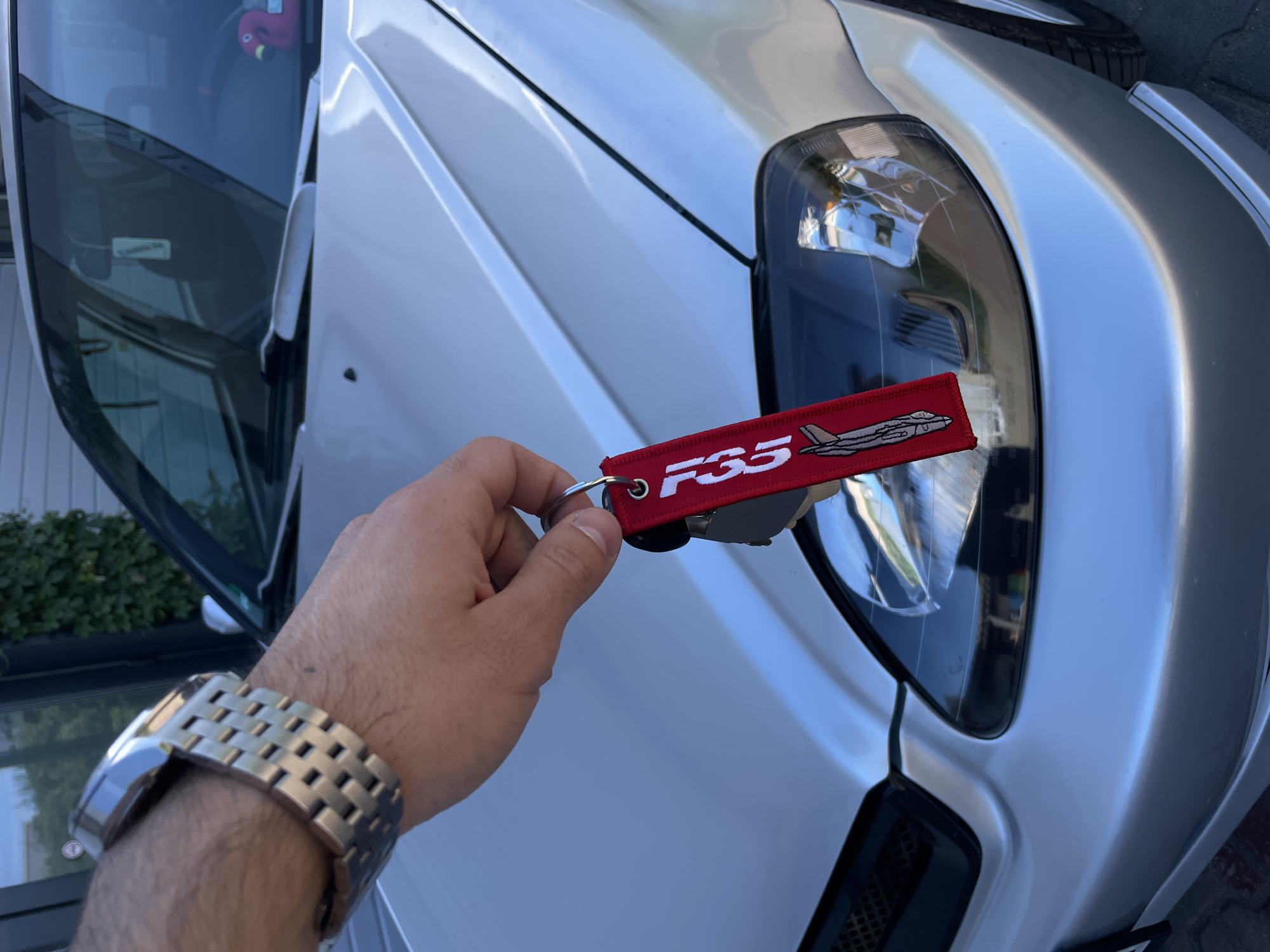 A close-up shot shows a person's hand holding a red keychain with the text "F35" and an image of a fighter jet. The keychain is positioned near the headlight of a silver car. The person is wearing a silver watch. The environment includes the car's body and headlight, as well as glimpses of foliage and what appears to be a wall. The location and city are unknown.