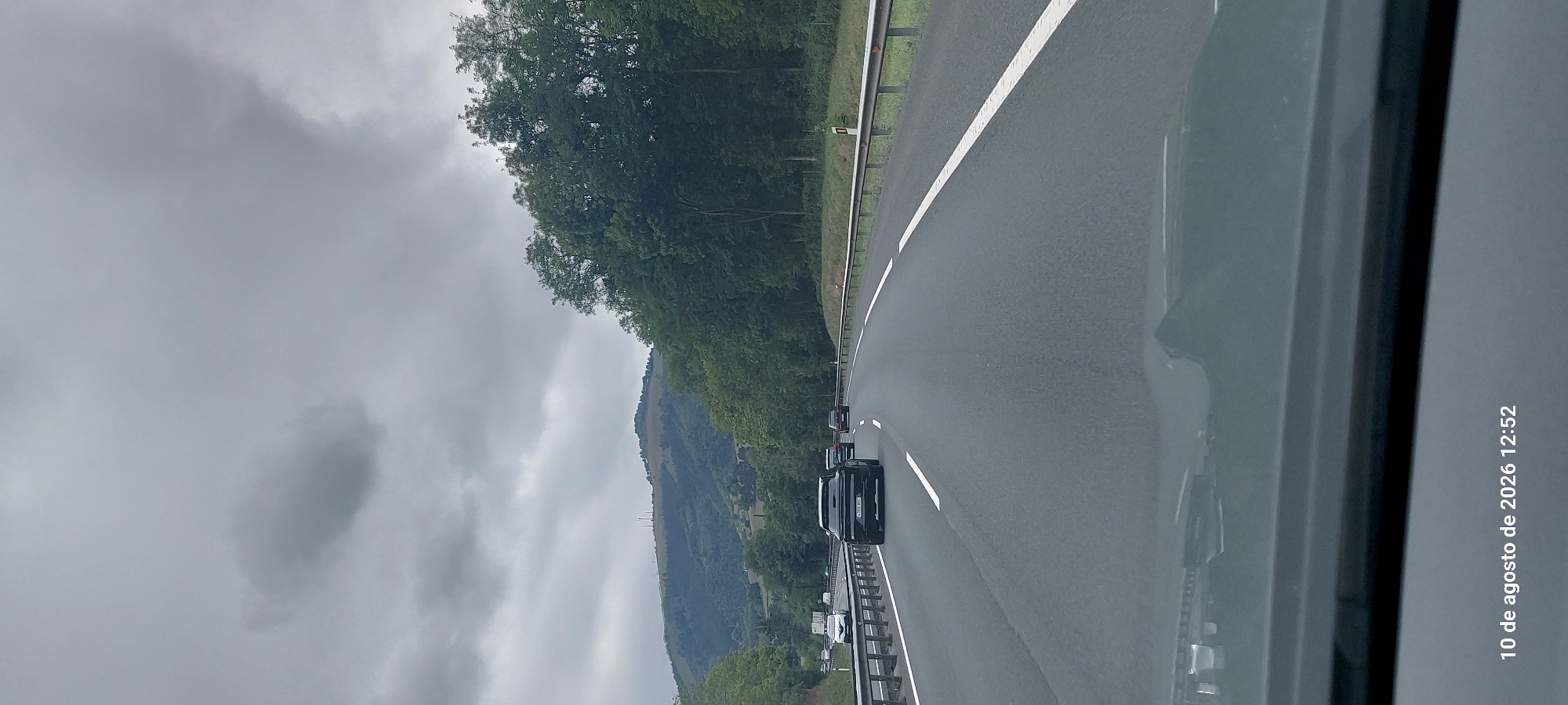 The image depicts a view from inside a vehicle traveling on a highway. The highway has multiple lanes and is bordered by a metal guardrail on the right. On the left side of the highway, there are trees and vegetation. In the background, a hilly landscape is visible with trees and some distant structures. The sky is overcast with grey clouds. The timestamp "10 de agosto de 2026 12:52" is visible in the lower left corner of the image, indicating the date and time the photo was taken. Several vehicles are present on the highway, including a black SUV in the foreground, a white van and other cars further ahead. The location is identified as Zestoa, Spain.