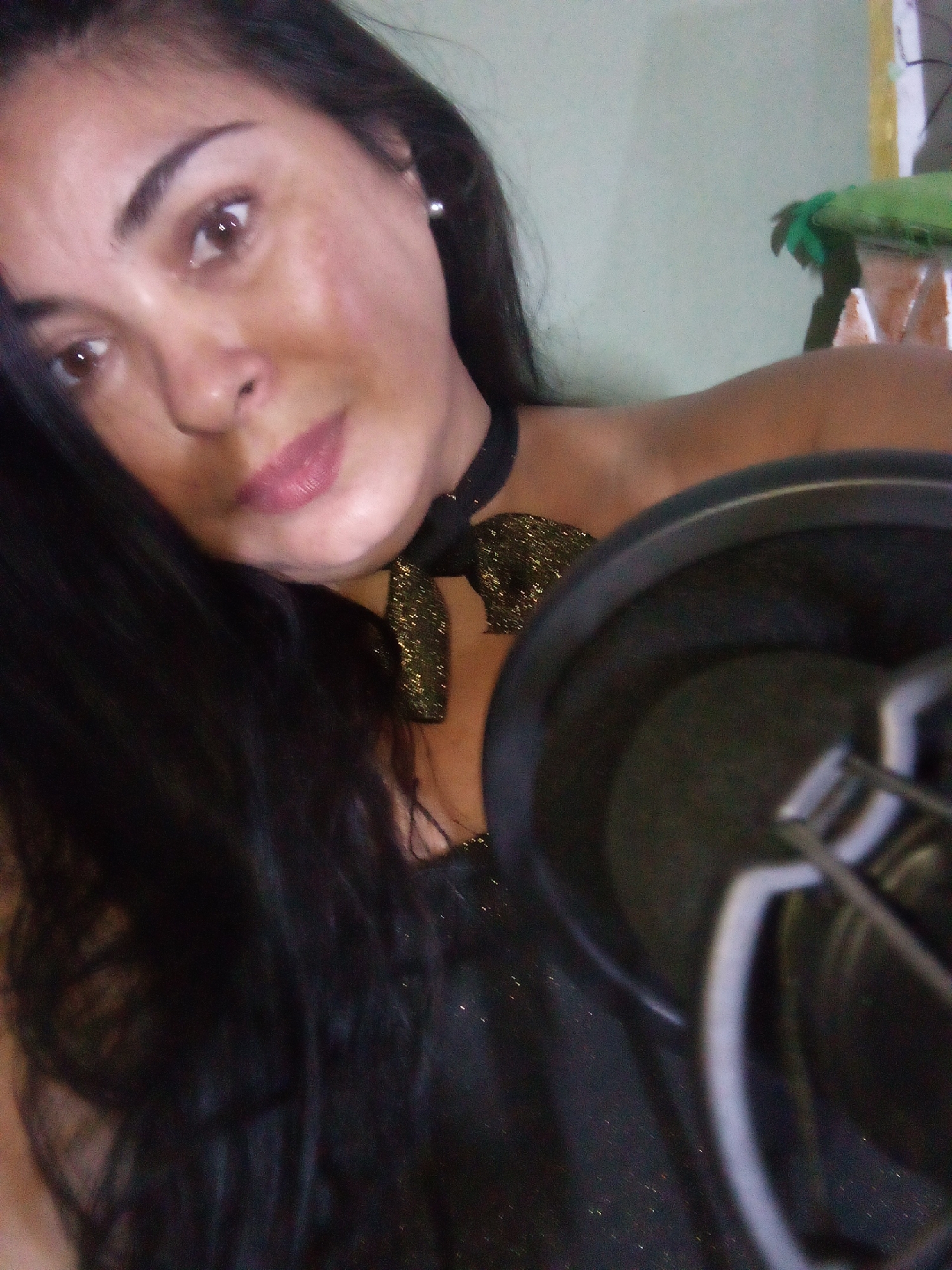 A medium shot captures a woman with dark hair leaning towards a microphone. She has brown eyes and pink lips. She's wearing a black top with gold glitter and a matching bow choker. A microphone with a pop filter is positioned prominently in the foreground. In the blurred background, a green object rests on a wooden structure. The image appears to have been taken indoors. There is no information available to determine a precise location in Juan Griego, Venezuela.