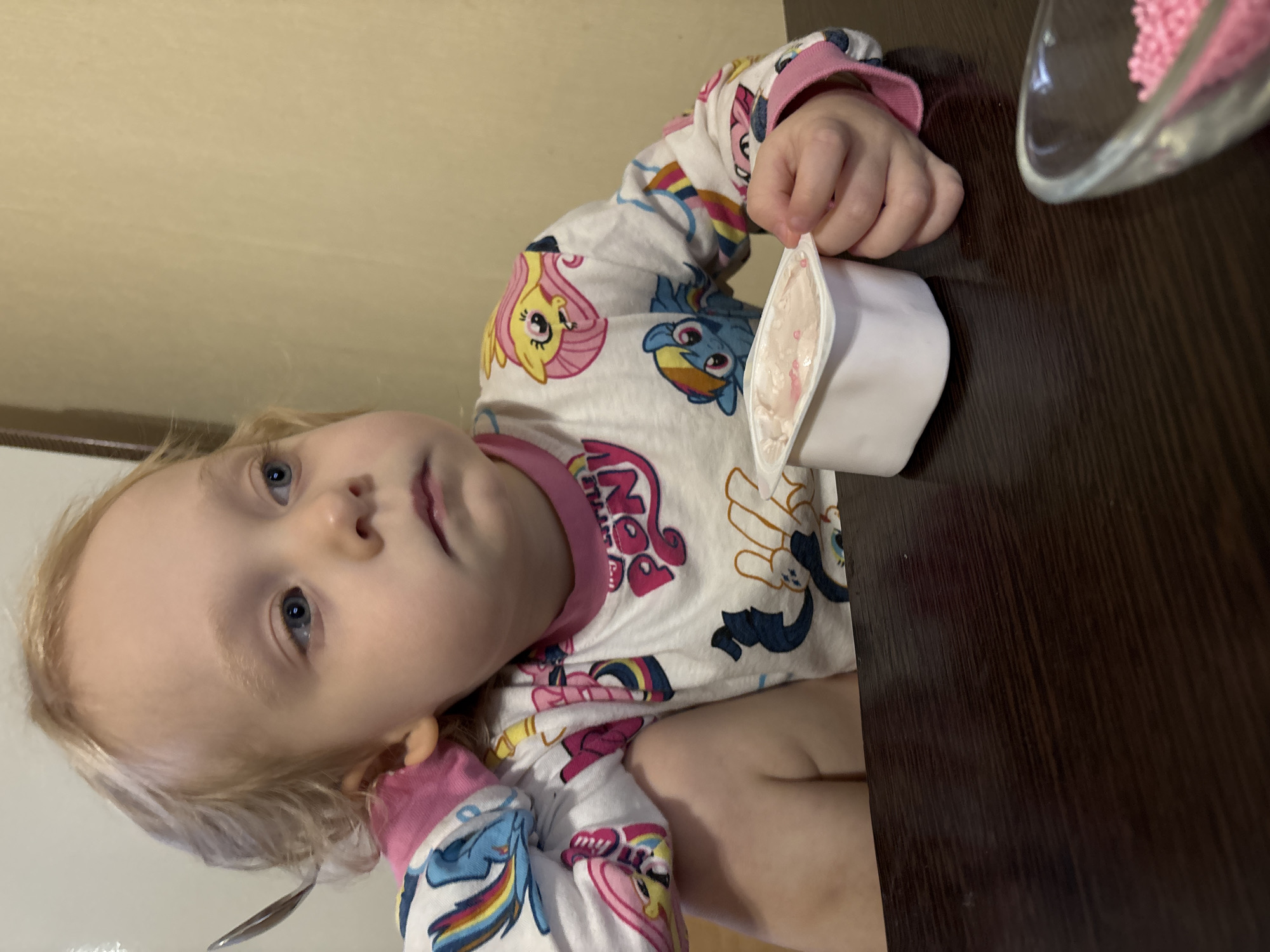 A toddler with blond hair and blue eyes, wearing a white long-sleeved shirt with "My Little Pony" characters, is sitting at a dark brown table. The child is holding a small white container of pink yogurt or cream. A clear glass bowl with pink sprinkles is also on the table. The background is a plain, light-colored wall. The location is Chaikovsky, Russia.