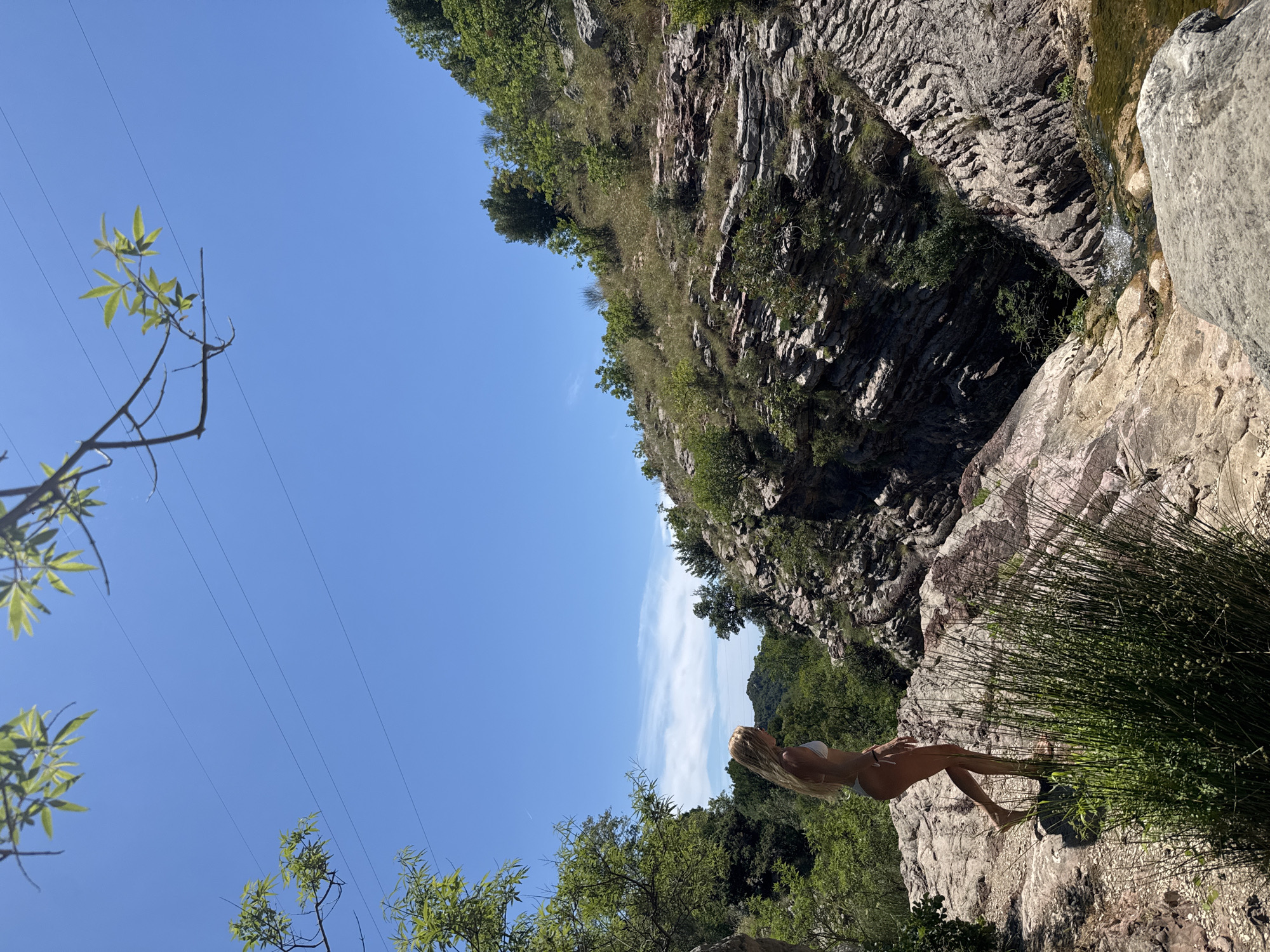 A blonde woman in a white bikini stands on a rocky outcrop overlooking a gorge. She is facing to her right and looking down into the gorge. The gorge has steep, rocky sides covered in vegetation. A small stream flows at the bottom of the gorge. The sky is clear and blue with a few wispy clouds. The scene is set in a natural, mountainous environment, likely in the hills surrounding Bečići, Montenegro.