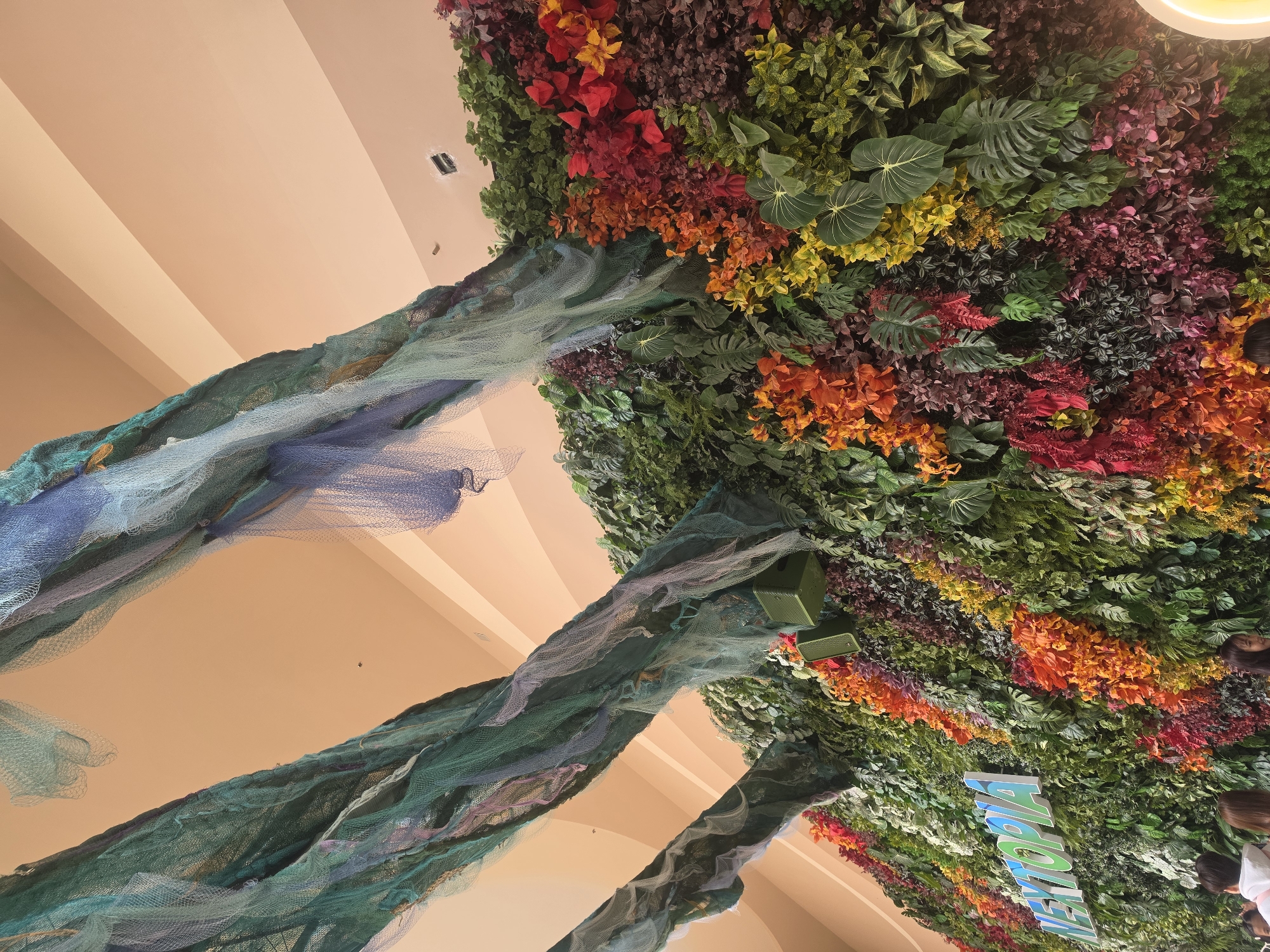 The image shows an indoor scene in Bangkok, Thailand, depicting a stylized natural setting. The scene is captured from a low angle, looking up towards a ceiling decorated to resemble a lush forest or garden. The ceiling is covered in a dense arrangement of artificial foliage, featuring a variety of colors, including green, red, orange, and yellow. The foliage appears to be a mix of leaves, flowers, and other plant elements.
In the foreground, there are structures resembling tree trunks or branches draped with netting of blue and green. These structures extend from the bottom of the frame towards the foliage-covered ceiling. In the lower right of the image, parts of a sign is visible. There is a blurred person and white shirt visible on the right edge of the frame. Overhead lighting is present.