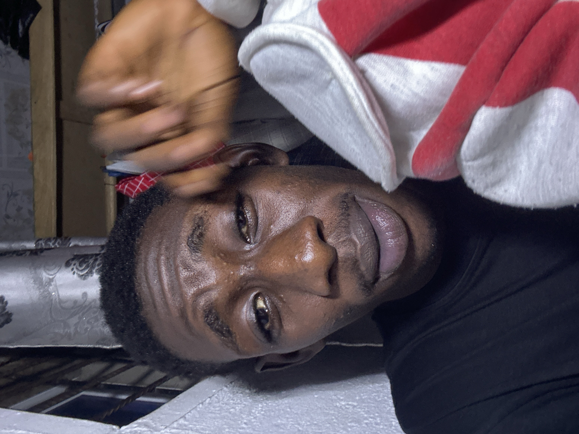 The image is a close-up selfie of a young Black male. He has dark skin, short, dark curly hair, and dark eyes. He is wearing a black t-shirt and appears to be indoors. His right hand, which is partially blurred, is raised in front of his face. Next to his face is a red and white cloth. The background shows a gray wall on the left and a curtain with a damask pattern behind him, with shelves and clothing visible above. There is no discernible location in the city of Durumi, Nigeria.