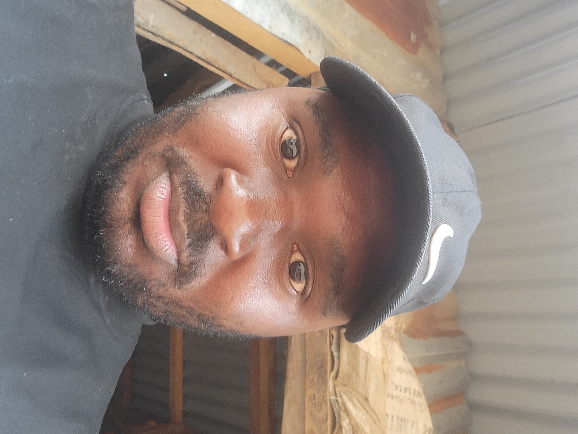 A close-up, eye-level selfie shows a Black man wearing a black t-shirt and a black baseball cap with a white Nike logo. He has brown eyes, a full beard, and a light-skinned complexion. The background is out of focus but appears to be a rustic, possibly unfinished structure with corrugated metal roofing and brown, textured walls, suggesting an outdoor or semi-outdoor setting in a developing area. The man is looking directly at the camera, and his expression is neutral. Based on the man's appearance and the visible background elements, this image is consistent with being taken in Madalla, Nigeria.