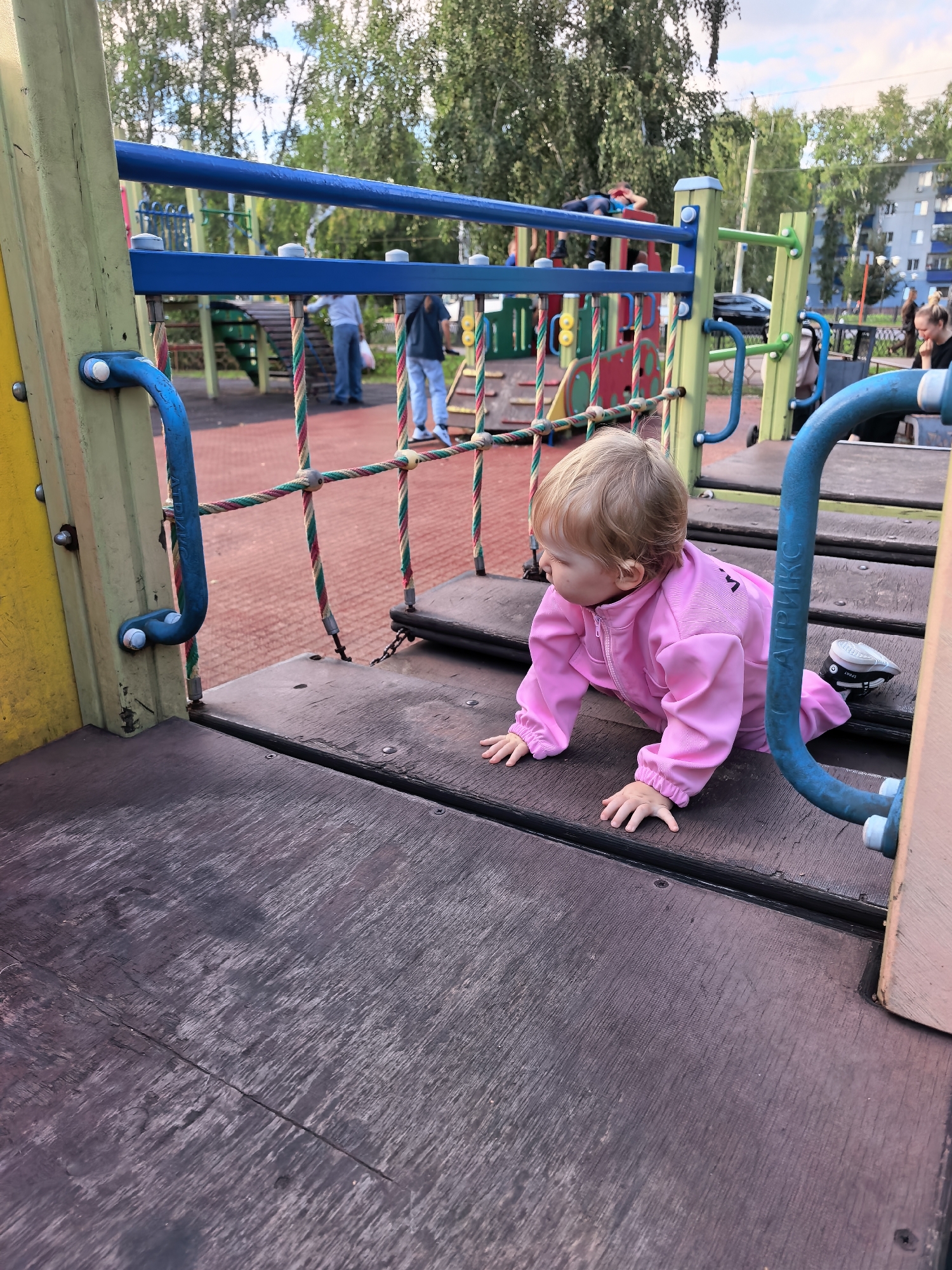 A young child in a pink tracksuit is crawling on a wooden platform of a playground. The child's back is to the camera and they are looking to the left. In the background, other children and adults are visible on the playground equipment. The playground is situated in a park-like setting with trees and buildings in the distance. The image appears to have been taken in the city of Sterlitamak, Russia.