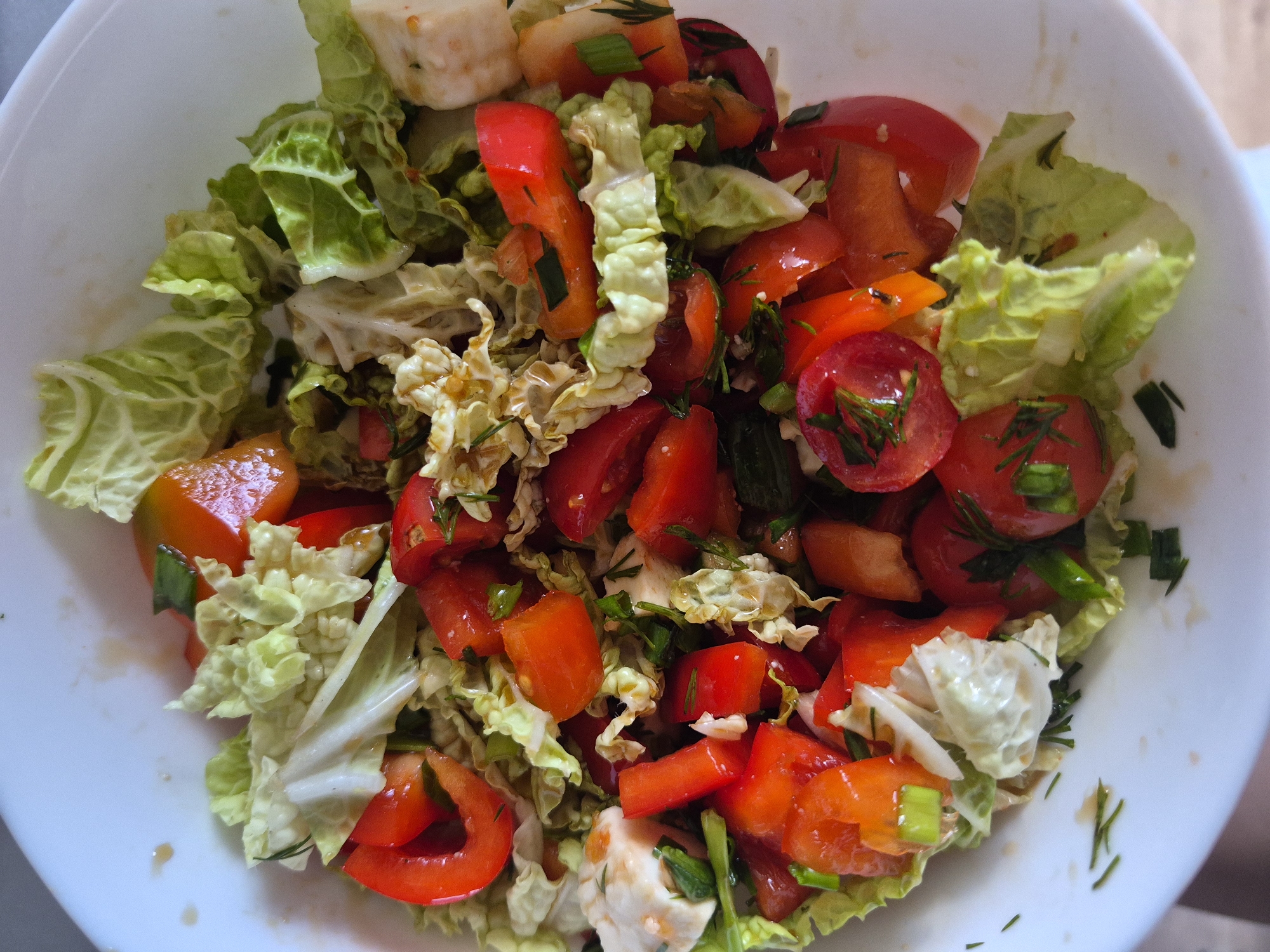 A top-down, close-up shot shows a white bowl filled with a colorful salad. The salad contains chopped red tomatoes, red bell peppers, green lettuce, white cheese cubes, and green herbs. The bowl appears to be sitting on a light-colored surface. The location is unknown.