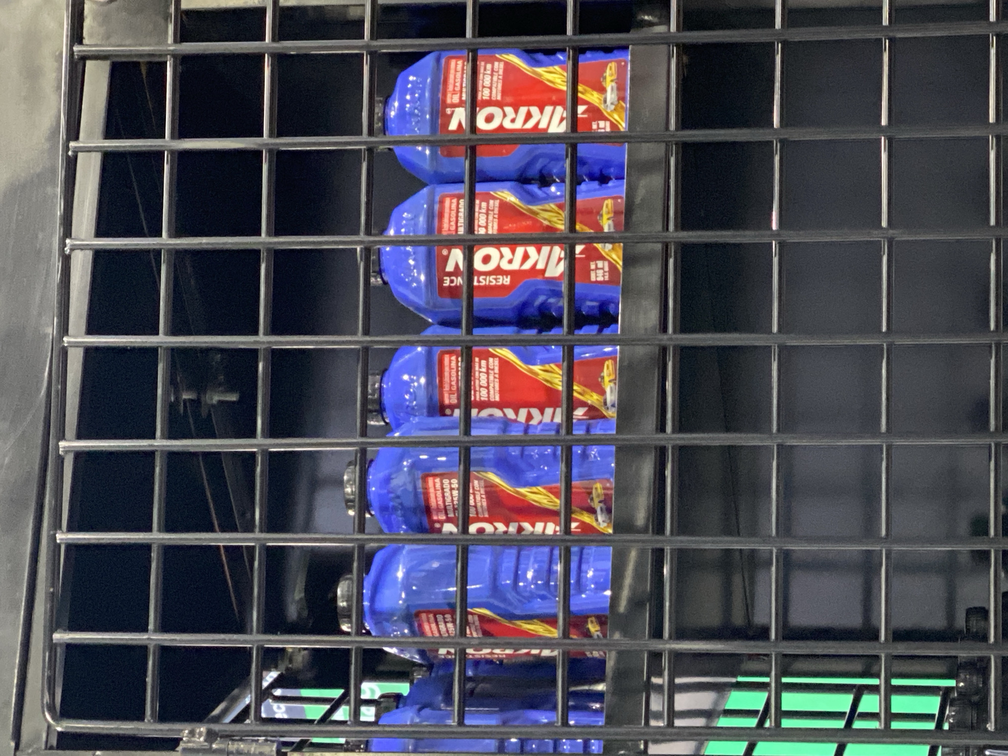 The image shows a stack of blue plastic containers labeled "KROV" behind a black wire rack. The containers appear to be motor oil, with labels indicating "GASOLINA," "ULTIGRADO," and "100,000 km." Some labels say "Resistence." The background is a mix of dark and light areas, with a glimpse of green at the bottom. The location is likely a store or storage area in Puebla, Mexico.