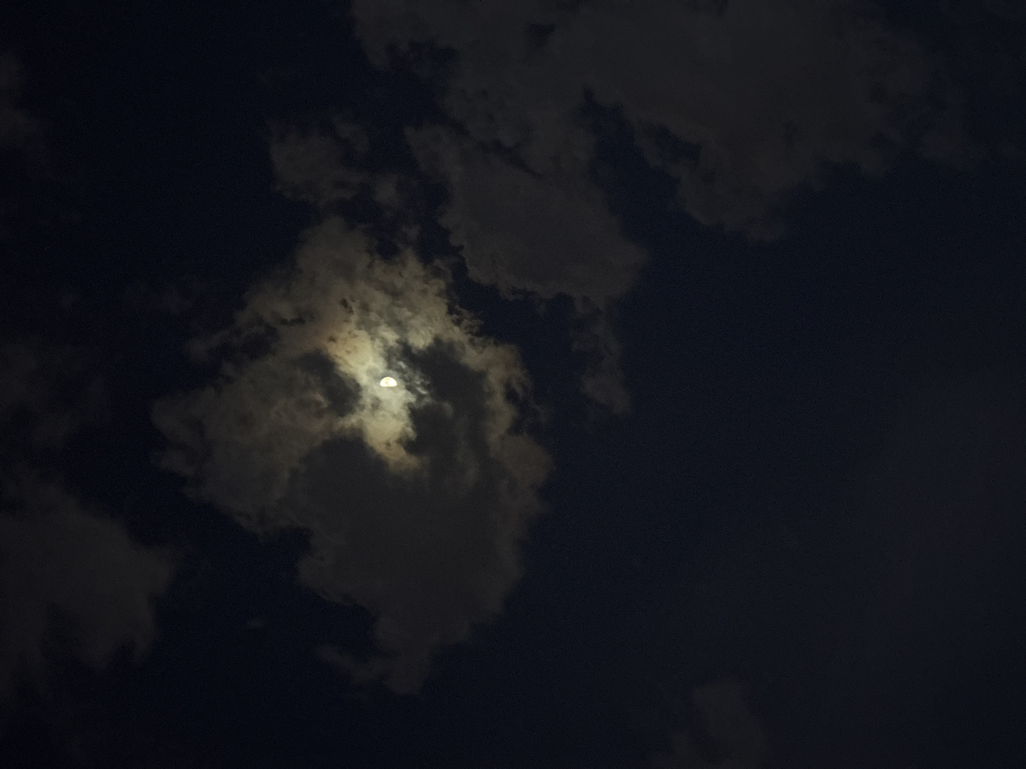 The image depicts a night scene in Bar, Montenegro. The sky is dark, with heavy cloud cover, and a partial moon is visible through a break in the clouds. No people, subjects, objects, actions, or interactions are visible other than the natural elements of the sky.