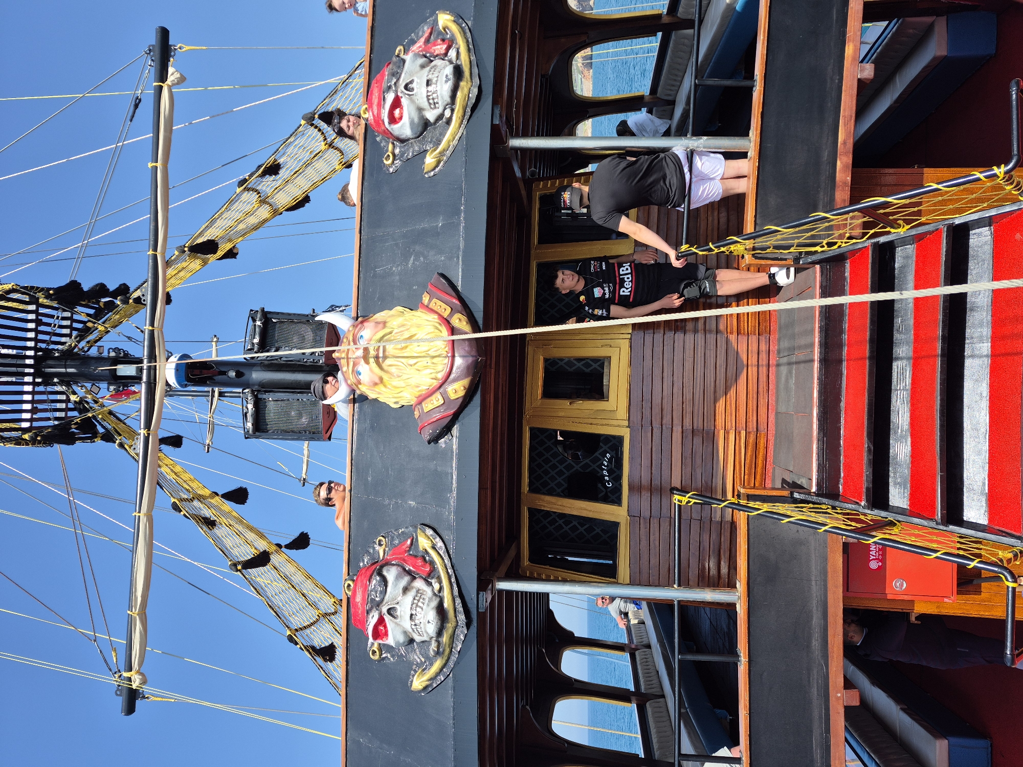 The image depicts a multi-level pirate-themed tourist boat, likely in Alanya, Turkey given the coastal setting and typical tourist attractions. The boat's exterior is adorned with pirate skull decorations and a figurehead. People are seen on different levels of the boat. On one level, two individuals are visible; one appears to be lying down, possibly relaxing, while the other is standing nearby. Other people are visible on higher decks, seemingly looking out at the view. The boat has masts, sails, and netting characteristic of a pirate ship replica. The weather appears sunny, as suggested by the bright blue sky. The boat is near the coast, with the sea visible through windows on the lower level.