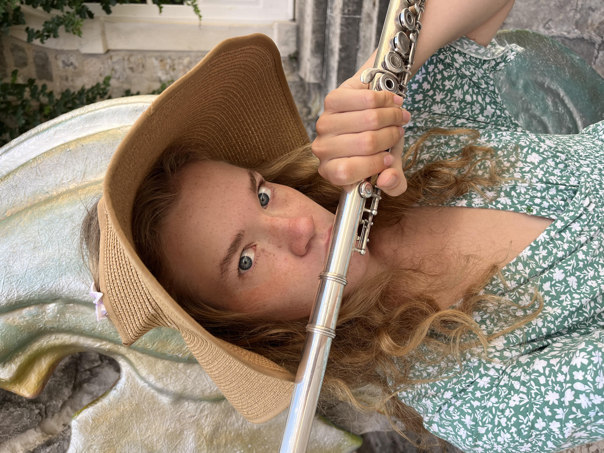 An angled, close-up portrait captures a young woman holding a silver flute in an outdoor setting in Budva, Montenegro. She has long, wavy reddish-blonde hair, striking blue eyes, and freckles across her nose and cheeks, and is looking directly at the camera with a wide-eyed expression. She wears a light brown straw hat or sun visor and a green dress with a white floral pattern. Her hands are positioned on the flute, and her mouth is near the mouthpiece, suggesting she is either playing or posing with the instrument.
The background shows natural light indicative of daytime. Behind her, elements of stone architecture are visible, partially covered by green foliage or vines. The woman appears to be leaning against or resting on a large, light-colored, textured object, possibly a sculpture or a decorative architectural feature, which takes up a significant portion of the lower and left side of the frame. The overall atmosphere is bright and engaging, showcasing a musical moment within a scenic environment. There is no visible text in the image.